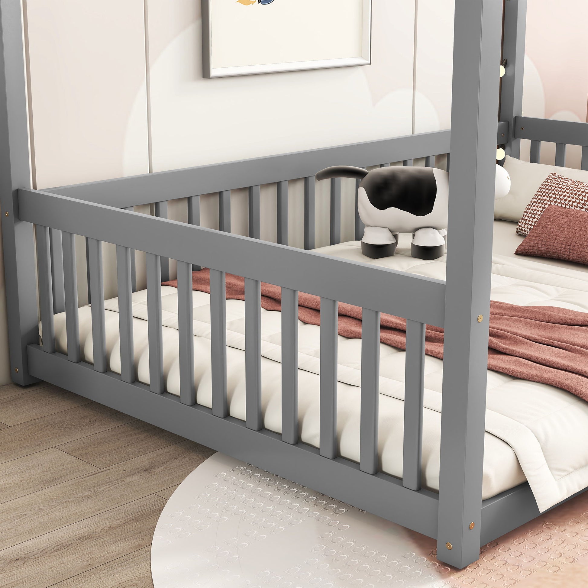 A Canopy Frame Floor Bed With Fence and guardrails in gray sits on a light wood floor. The bed has a white mattress, peach blanket, and pillow. A plush cow toy is near the head of the bed, with a round rug partially visible nearby.