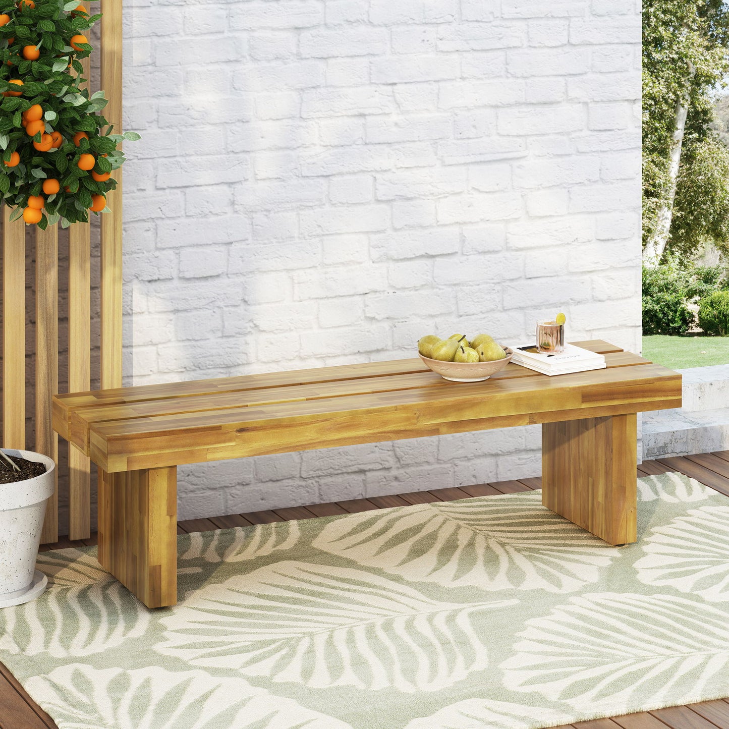 The Nahanni Rustic Acacia Wood Bench with Slat Design in Natural sits on a green and white leaf-patterned rug by a white brick wall, topped with a bowl of pears and juice. A potted orange tree and book complete the outdoor scene.