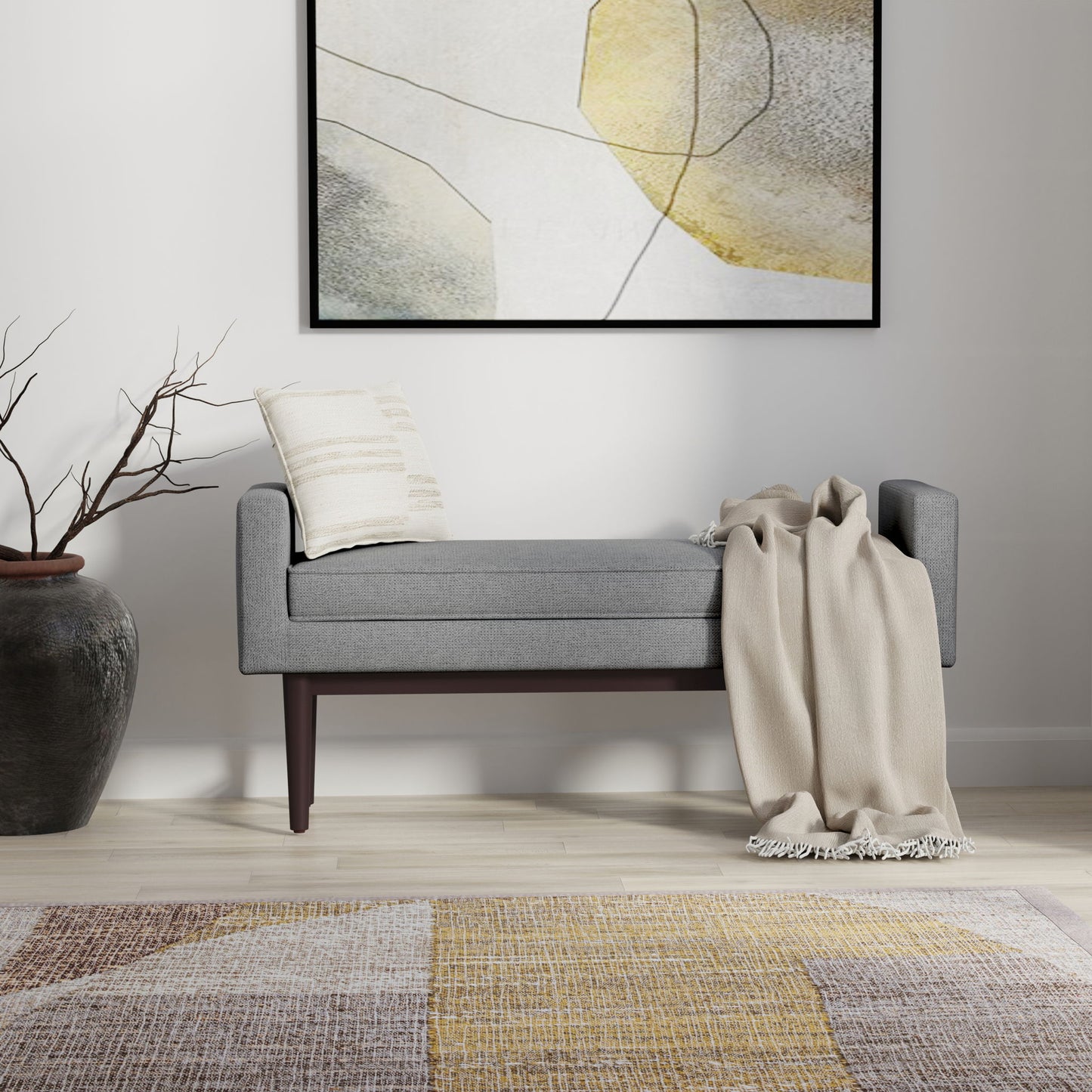 The Scott Upholstered Ottoman Bench in modern gray features dark wooden legs, topped with a striped pillow and beige throw. Above hangs abstract art, while a tall vase with branches and a patterned rug complete the stylish look.