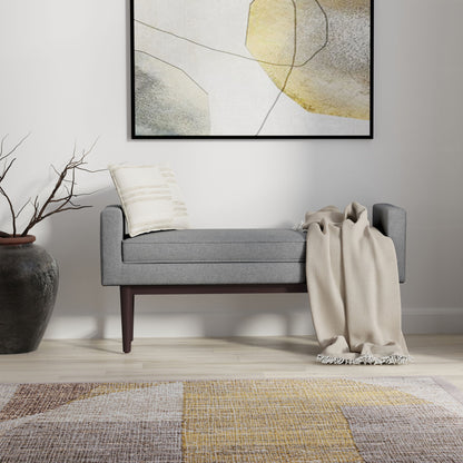 The Scott Upholstered Ottoman Bench in modern gray features dark wooden legs, topped with a striped pillow and beige throw. Above hangs abstract art, while a tall vase with branches and a patterned rug complete the stylish look.