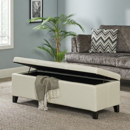 The Glouster Contemporary Storage Ottoman, shown in cream with its lid open to reveal a gray fringed blanket inside, sits on a beige rug in a living room with a gray sofa, chevron pillow, potted plant, and window.