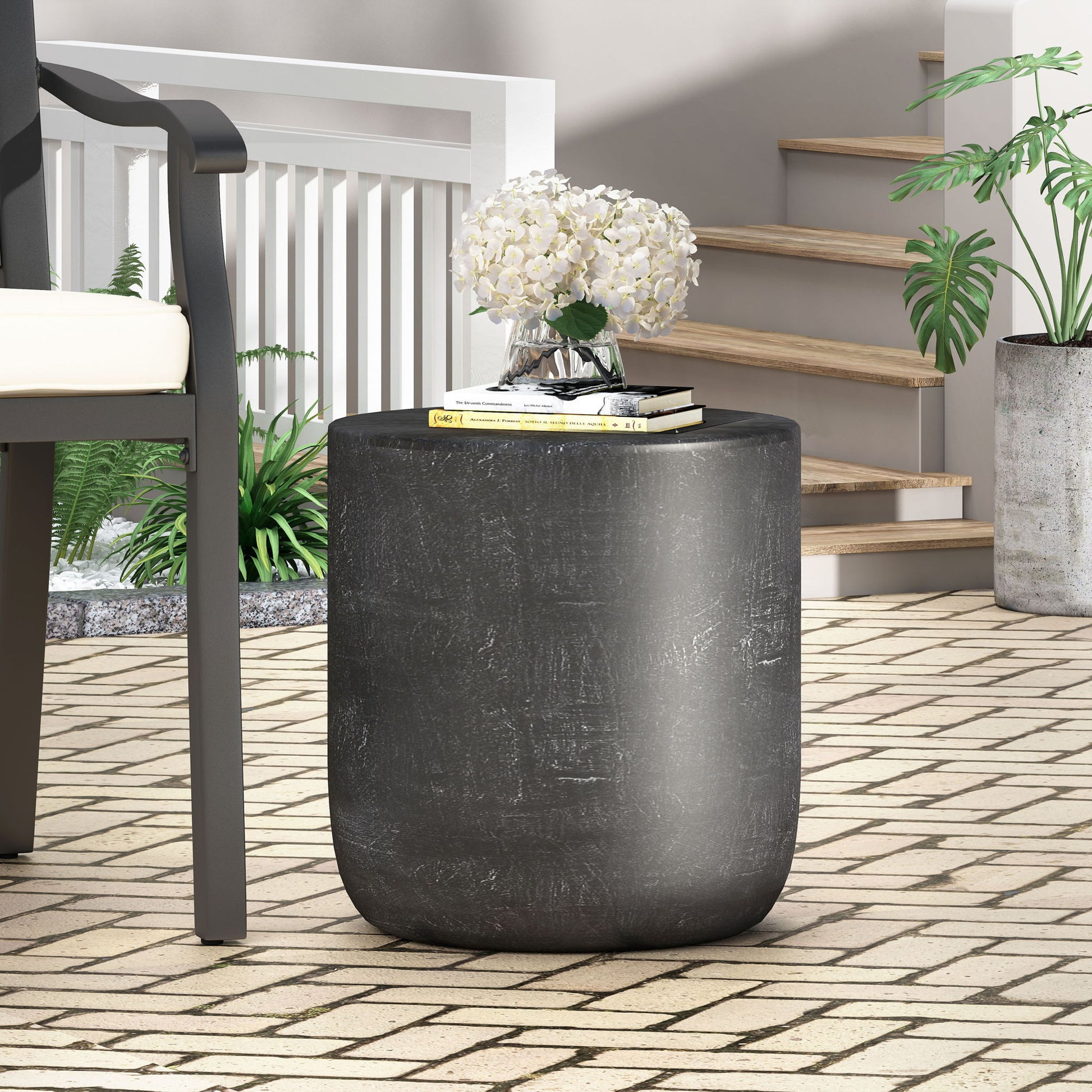 The Outdoor Lightweight Concrete End Table With Natural Texture - Matte Black sits on a tiled patio by a white railing, alongside a potted plant and wooden stairs, topped with white flowers, books, and glasses.