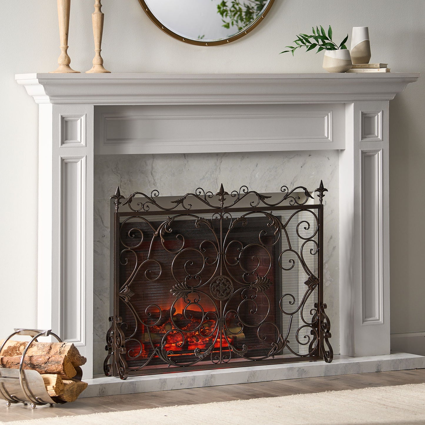 A white fireplace features the Darcie - Wrought Fireplace Screen with an ornate design, glowing embers inside, a round mirror and candles on the mantel, and a log holder with firewood beside it.