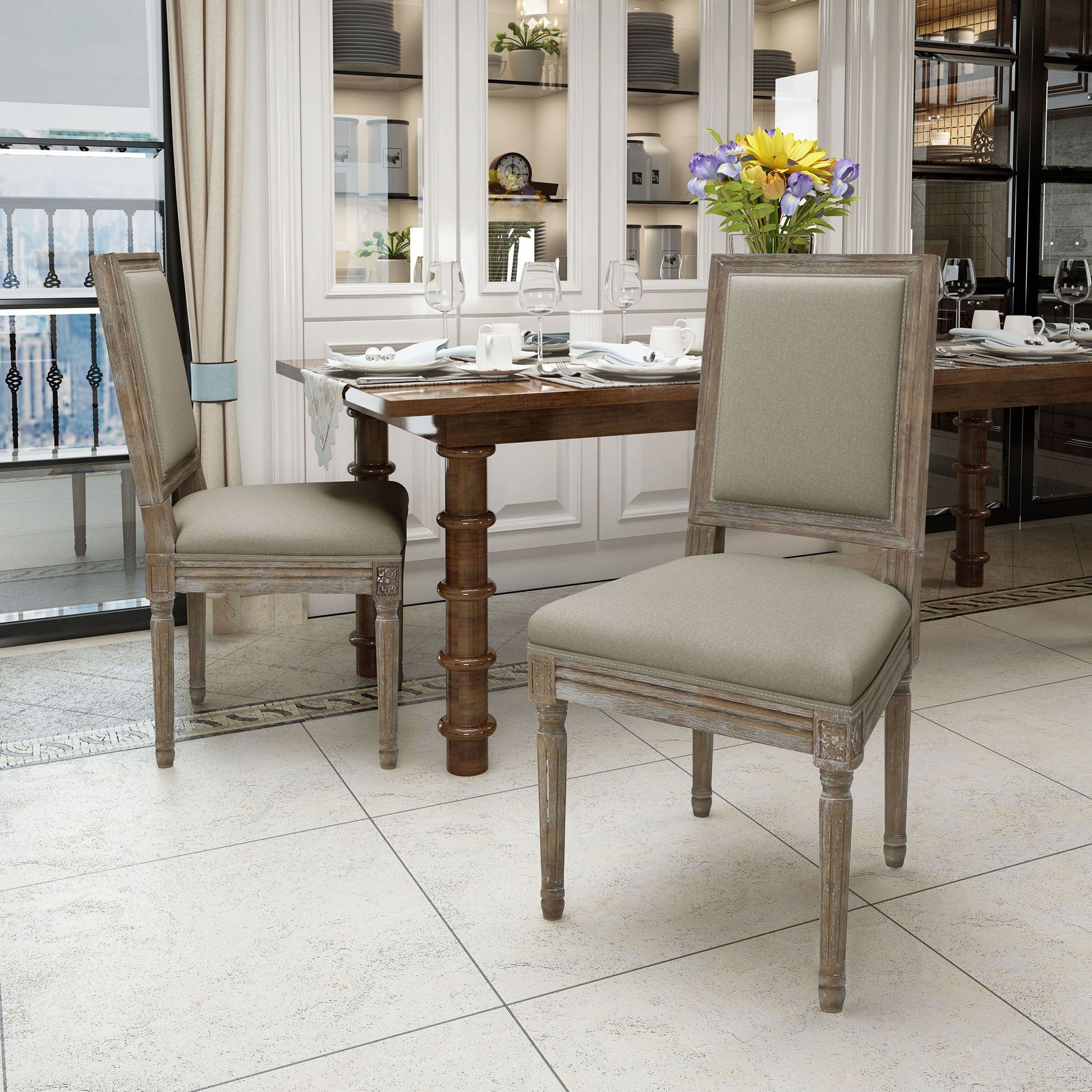 Two Traditional Style Dining Chairs With Fabric Upholstery and wooden frames sit near a set dining table with plates and glasses. The room has tiled floors, large windows, cabinets, and a vase of flowers on the table.