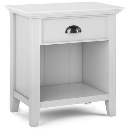 The Acadian Handcrafted Bedside Table is a white wooden nightstand with a single drawer, metal handle, open lower shelf, and four sturdy legs.