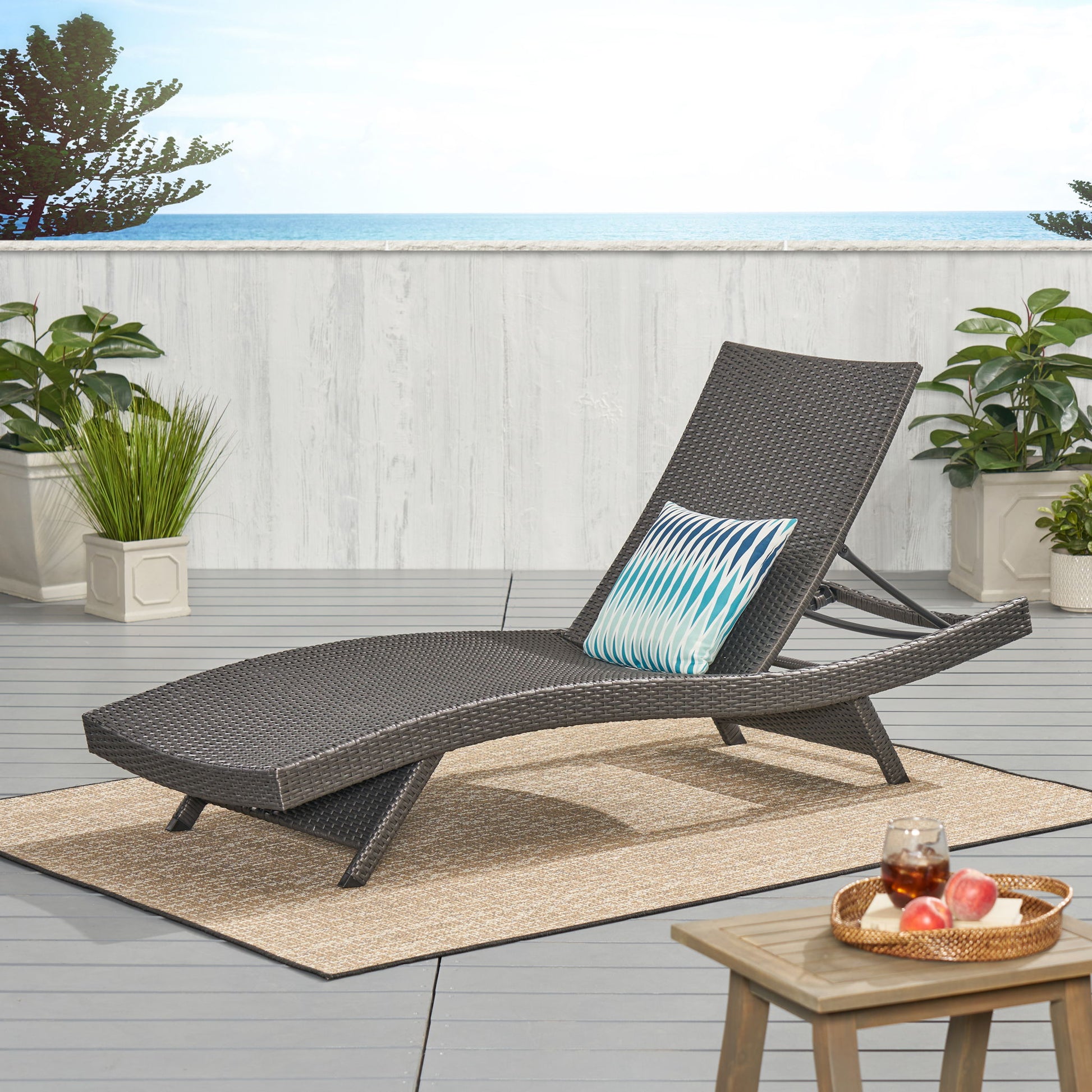 The Salem - Chaise Lounge with a blue patterned pillow rests on a patio rug amid green plants. A small table holds a tray with drinks and fruit, while the ocean and blue sky form the scenic backdrop.