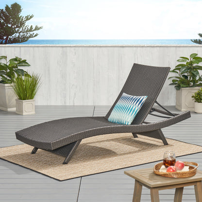 The Salem - Chaise Lounge with a blue patterned pillow rests on a patio rug amid green plants. A small table holds a tray with drinks and fruit, while the ocean and blue sky form the scenic backdrop.