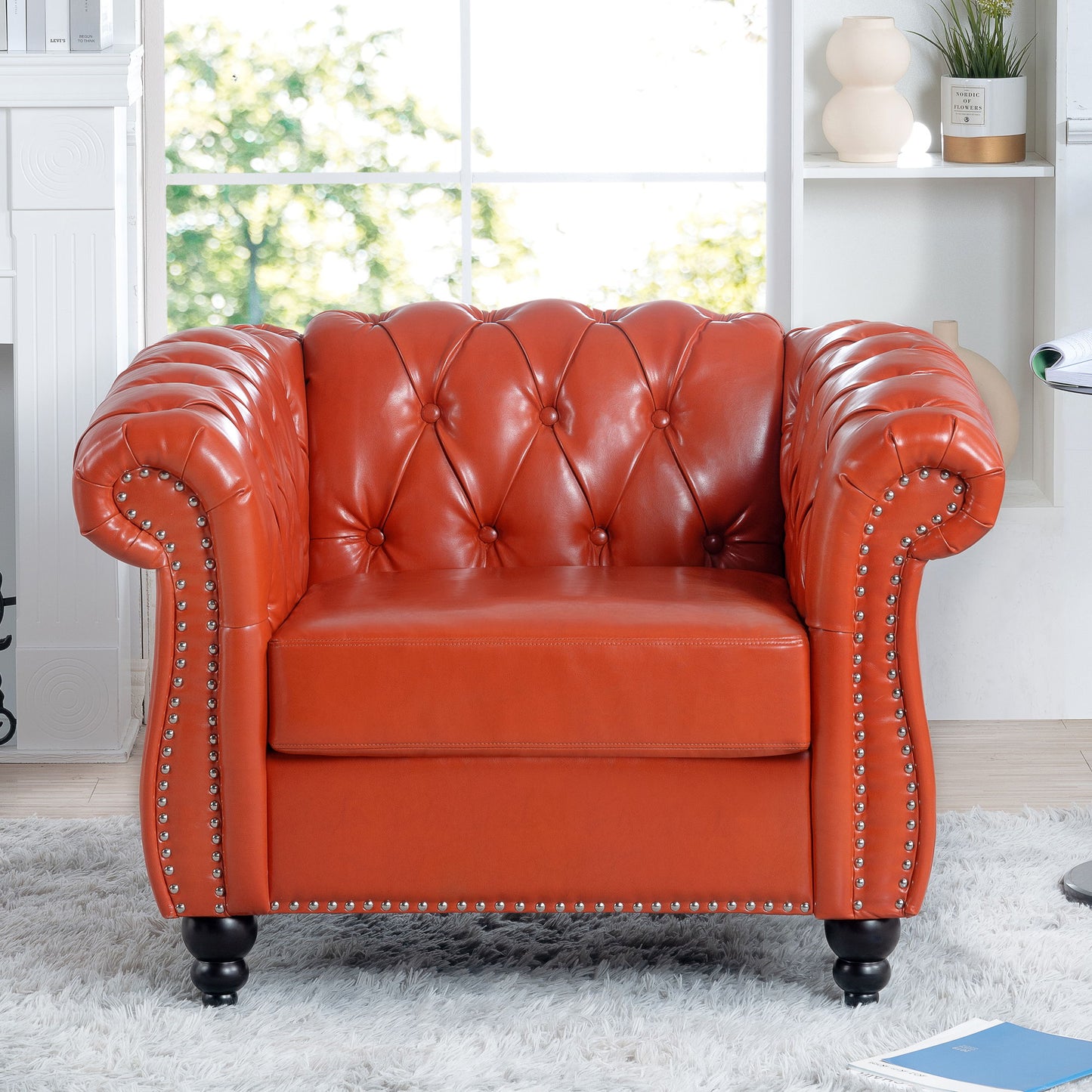A 1 Seater Sofa For Living Room in red leather with button tufting and silver nailhead trim is placed on a white shag rug in a modern, bright living room with shelves and large windows in the background.
