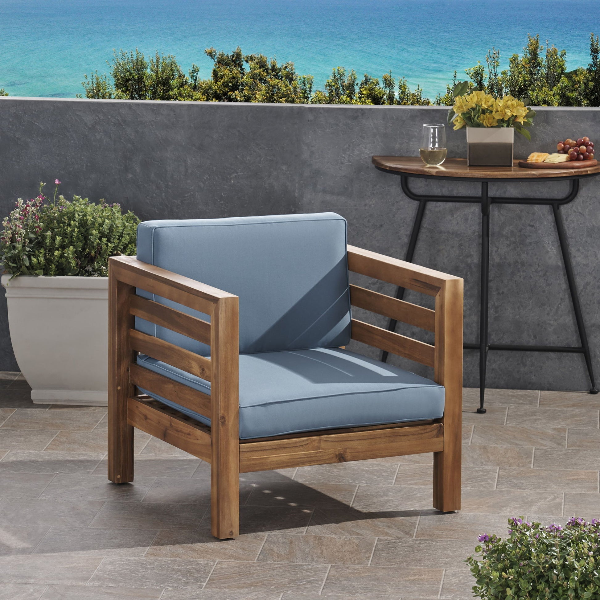 An Outdoor Acacia Wood Patio Chair With Fabric Cushion in light blue sits on a tiled terrace, with a planter of green plants, a table set with food and drink, and a scenic view of trees and the blue ocean in the background.