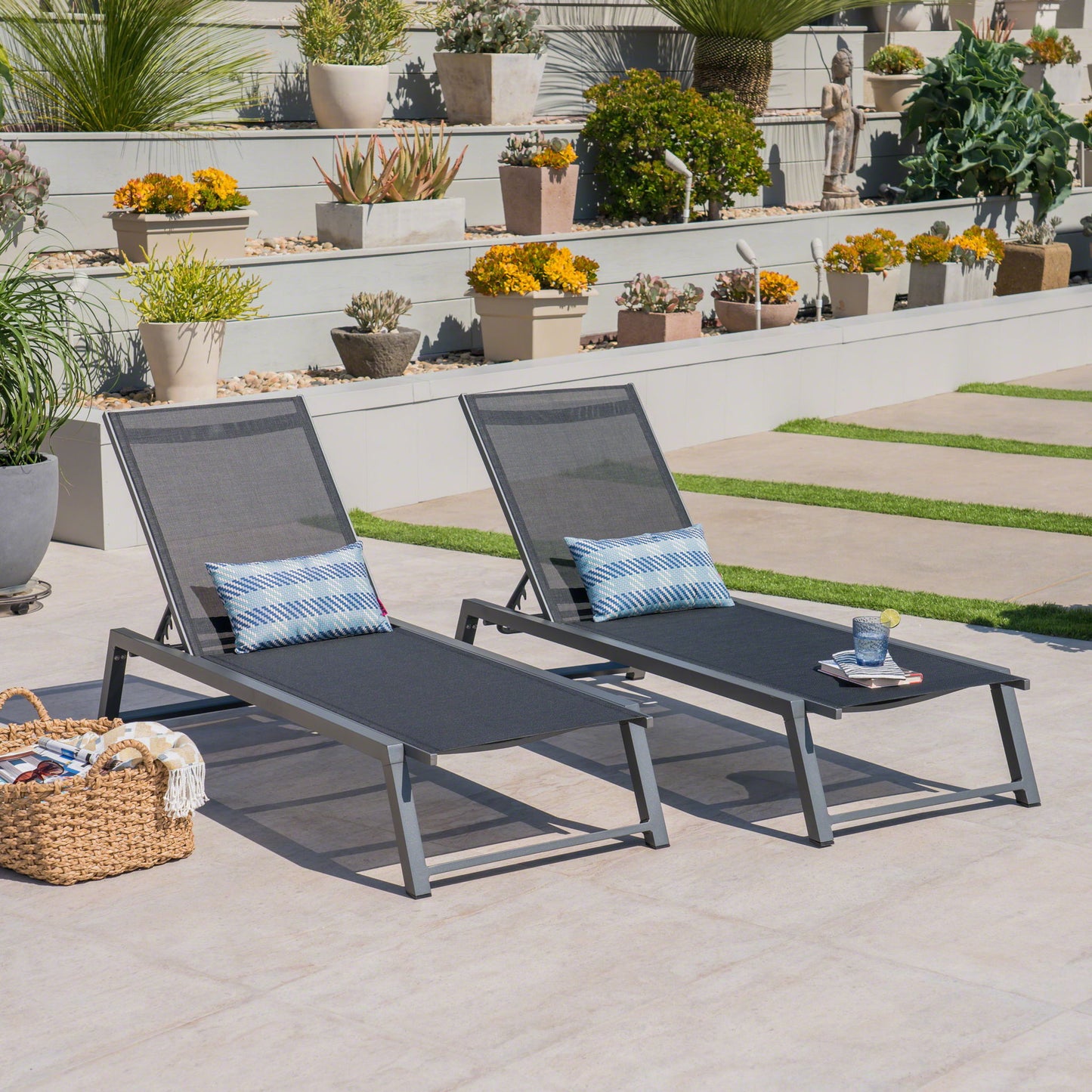 Two Myers Modern Chaise Lounge chairs with adjustable mesh backs sit on a tiled patio with blue patterned pillows and a wicker basket, surrounded by potted plants and flowers on sunny tiered steps.