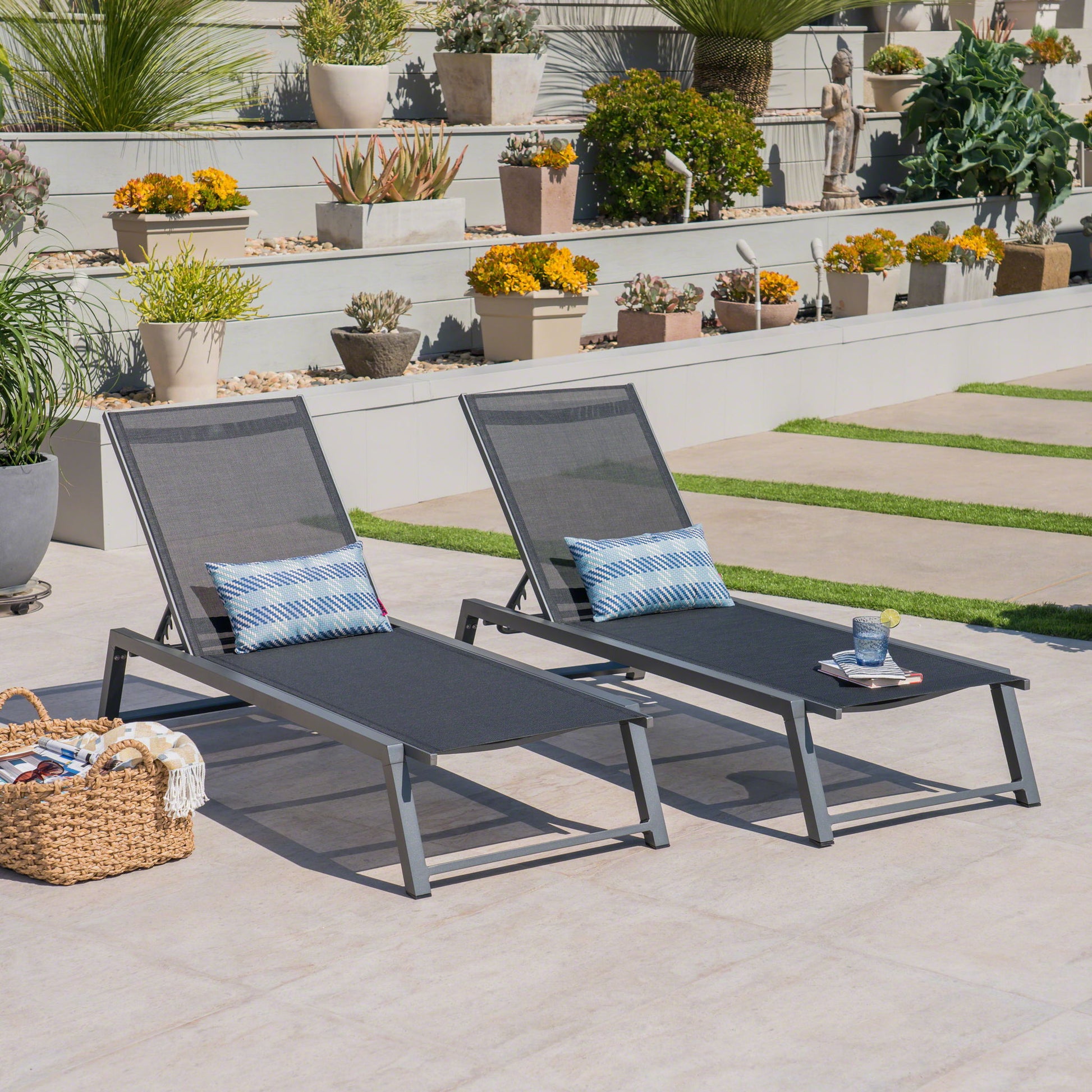 Two Myers Modern Chaise Lounge chairs with adjustable mesh backs sit on a tiled patio with blue patterned pillows and a wicker basket, surrounded by potted plants and flowers on sunny tiered steps.