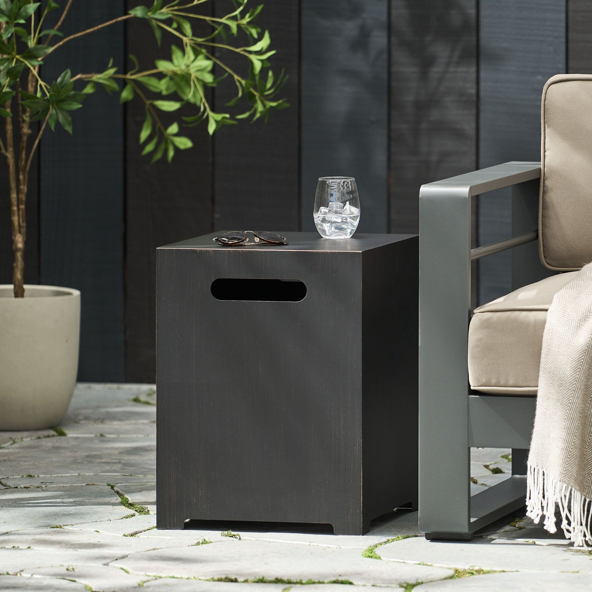 A modern outdoor scene featuring the Modern Tank Holder Side Table With Powder Coated Frame, displaying eyeglasses and a glass of water beside a beige cushioned chair with a throw, on a stone patio with a potted plant nearby.
