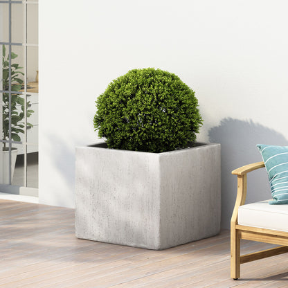 A round green shrub in a large Modern Outdoor Planter sits on a wooden deck beside a white outdoor sofa with green and white striped pillows, with indoor plants visible through the glass door in the background.