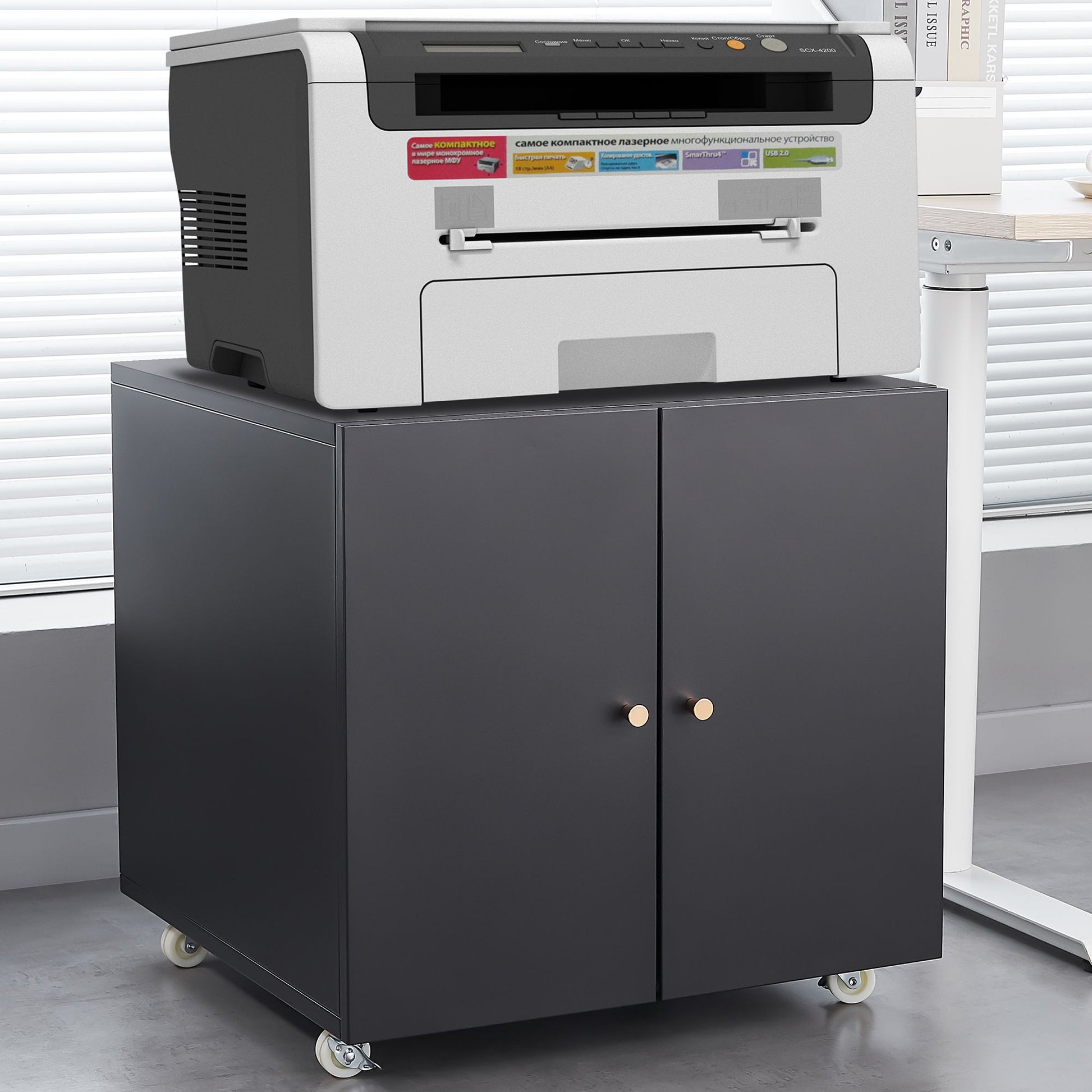 An Office Furniture Copier Cabinet 2 Door Steel Copier Stand Mobile Pedestal File Printer Stand holds a gray and black multifunction printer, standing on caster wheels in an office with light-colored blinds behind it.