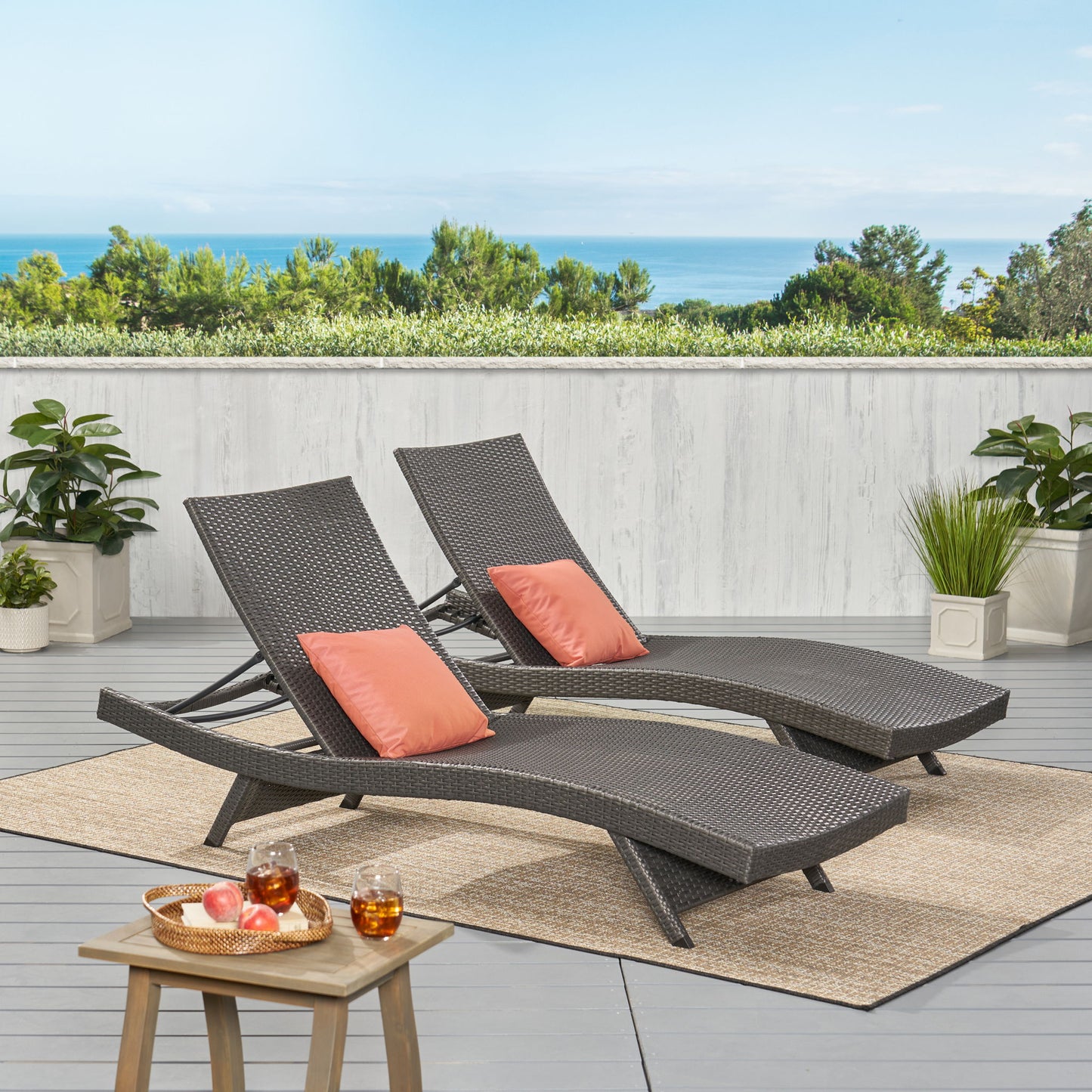 Two Salem - Chaise Lounges with coral pillows rest on a woven rug on the patio, surrounded by potted plants. A small table holds drinks and fruit. A white fence, lush greenery, and an ocean view complete the serene setting.