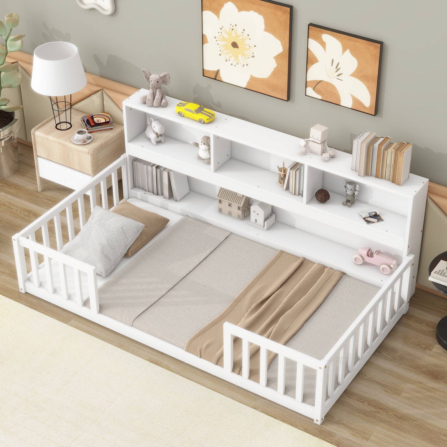 A tidy children's bedroom with a white Floor Bed With Bedside Bookcase, featuring guardrails, beige bedding, headboard shelves for toys and books, plus a small table, lamp, and wall art nearby.