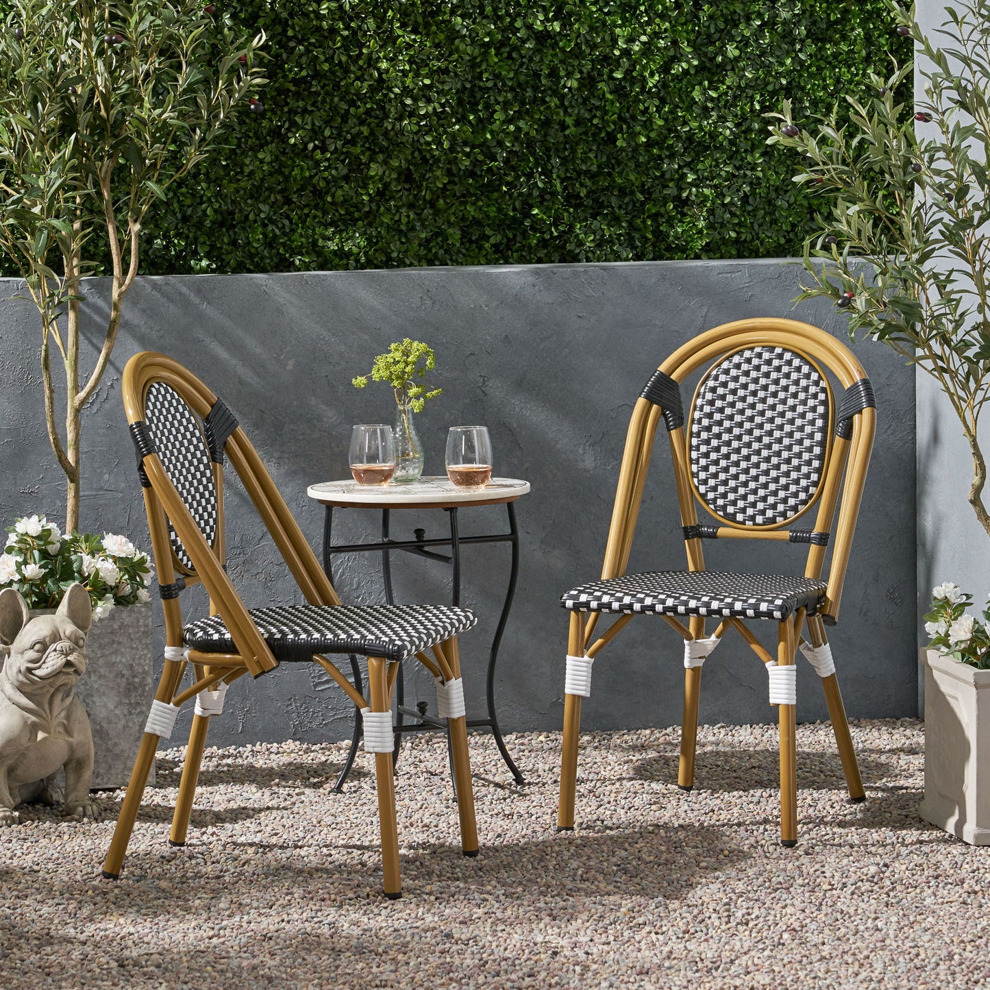 Two Outdoor Rattan And Aluminum French Bistro Chairs with black-and-white patterned seats sit at a small round table holding two drinks and a vase, surrounded by plants, a stone statue, and a gray wall with green foliage above.
