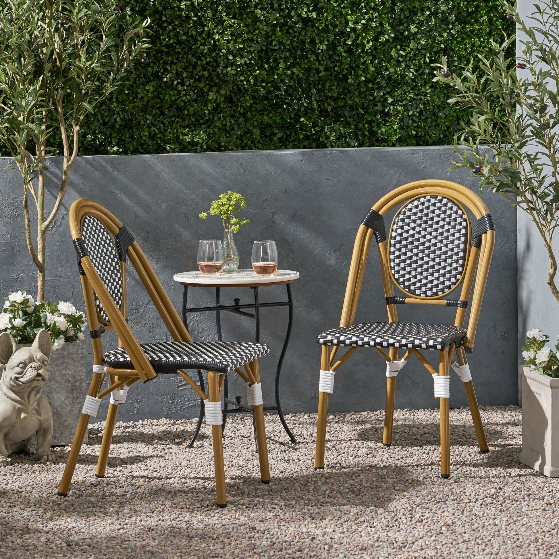 Two Outdoor Rattan And Aluminum French Bistro Chairs with black-and-white patterned seats sit at a small round table holding two drinks and a vase, surrounded by plants, a stone statue, and a gray wall with green foliage above.