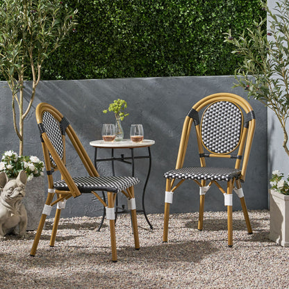 Two Outdoor Rattan And Aluminum French Bistro Chairs with black-and-white patterned seats sit at a small round table holding two drinks and a vase, surrounded by plants, a stone statue, and a gray wall with green foliage above.