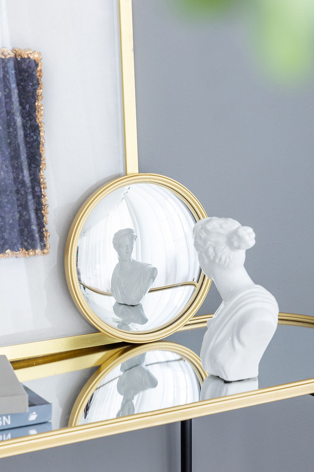 A white bust sits on a glass table with a gold rim, reflecting in the Round Mirror—Circle Mirror With Iron Frame—for living room, bedroom, vanity, entryway, or hallway. Modern wall art decorates the gray wall in the background.