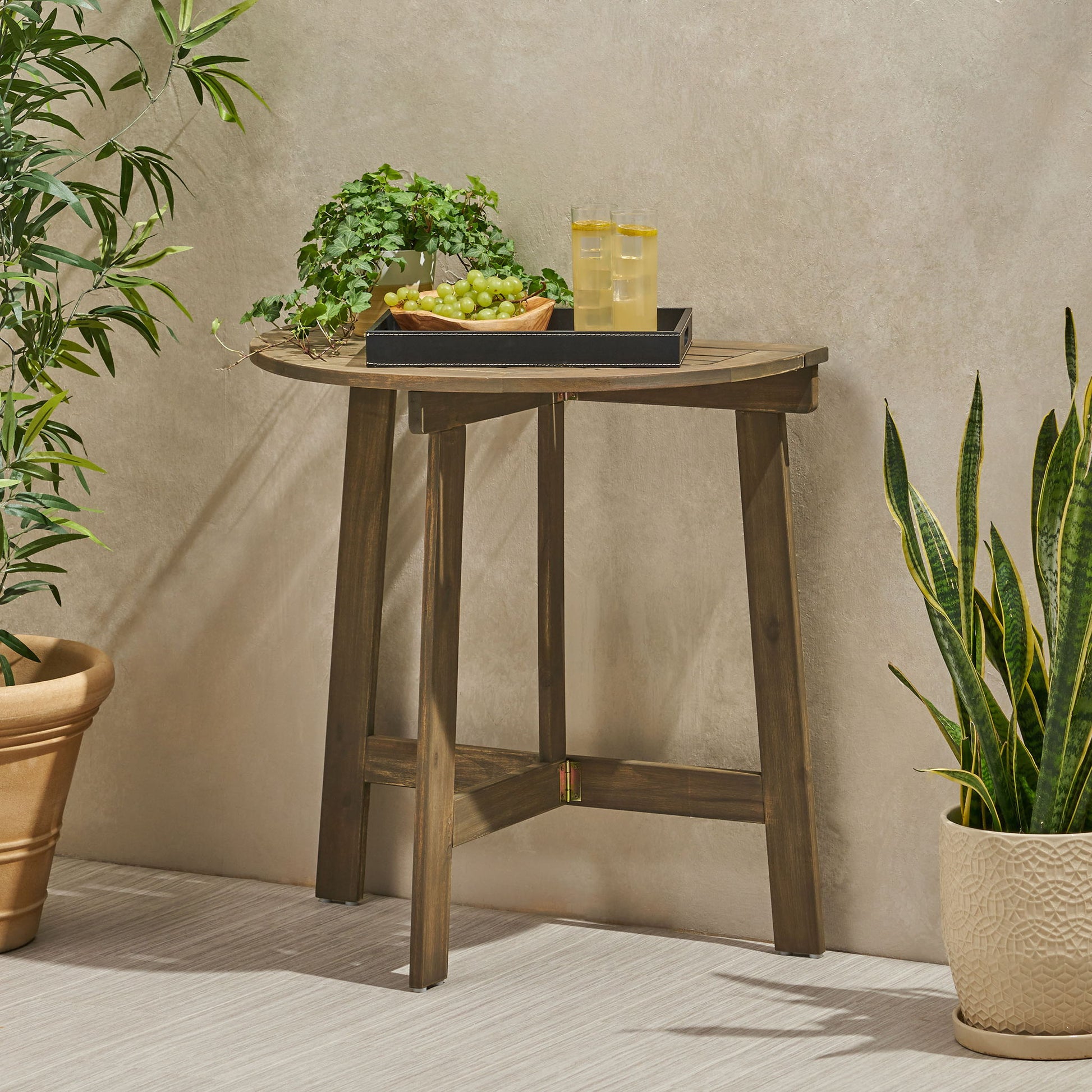 An Outdoor Half Round Folding Acacia Wood Bistro Table holds a black tray with two glasses of lemonade, a bowl of green grapes, and a potted plant. The table sits against a beige wall beside two more potted plants.