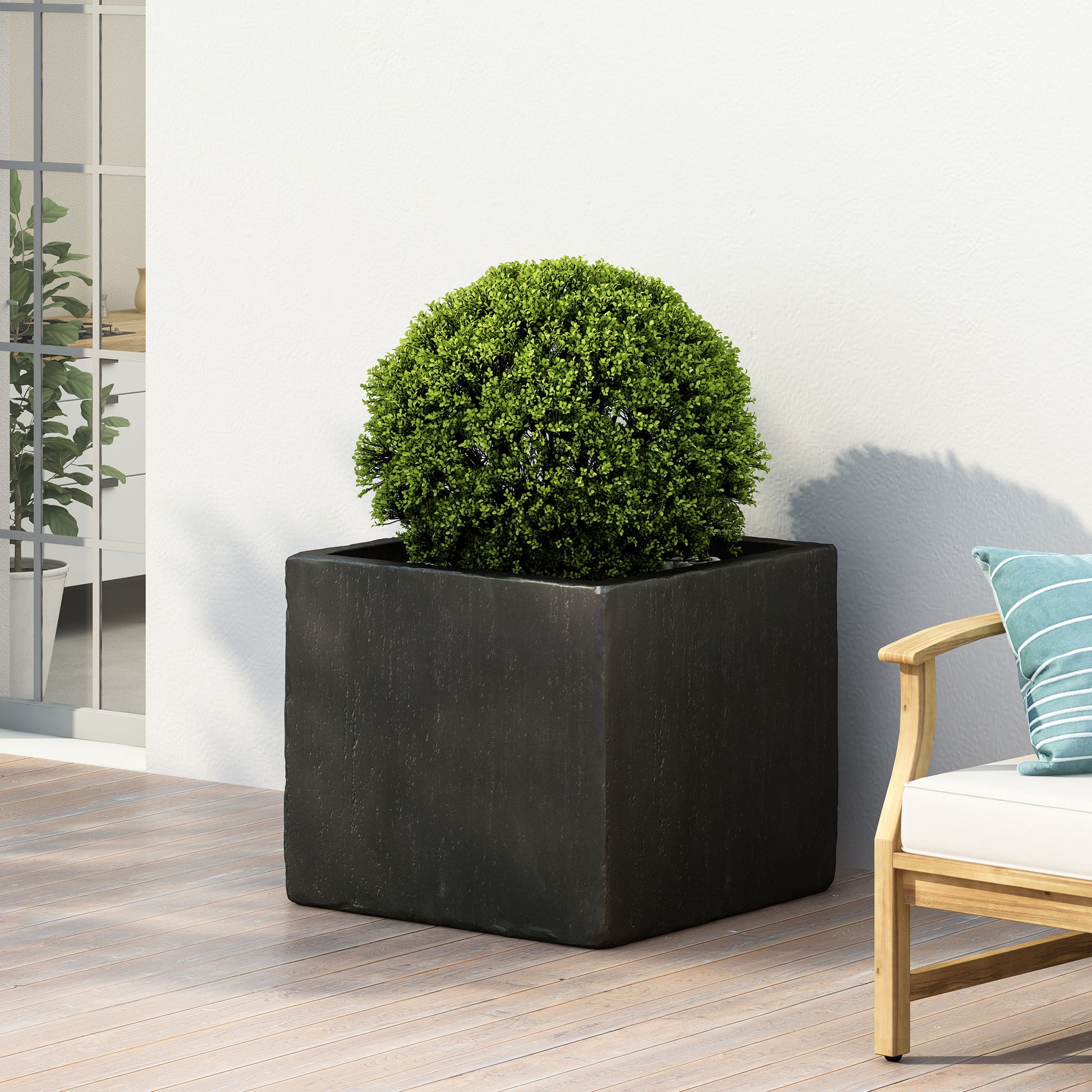 A round green shrub in a Modern Outdoor Planter sits on a wooden patio beside a white outdoor sofa with blue and white striped cushions, against a white wall.