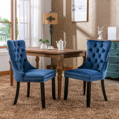 Two Nikki modern tufted velvet dining chairs with nailhead trim stand on a brown rug before a wooden table set with a teapot in a cozy room featuring large windows, framed art, and decorative decor.
