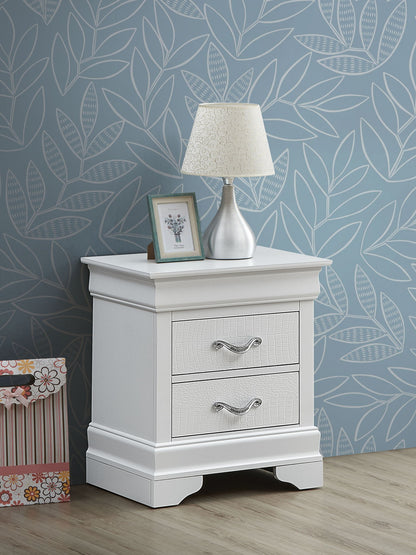 The Lorana Nightstand, featuring two drawers with silver handles, stands on a wooden floor. On top are a silver lamp with a patterned shade and a framed photo. Blue leaf-patterned wallpaper decorates the background.