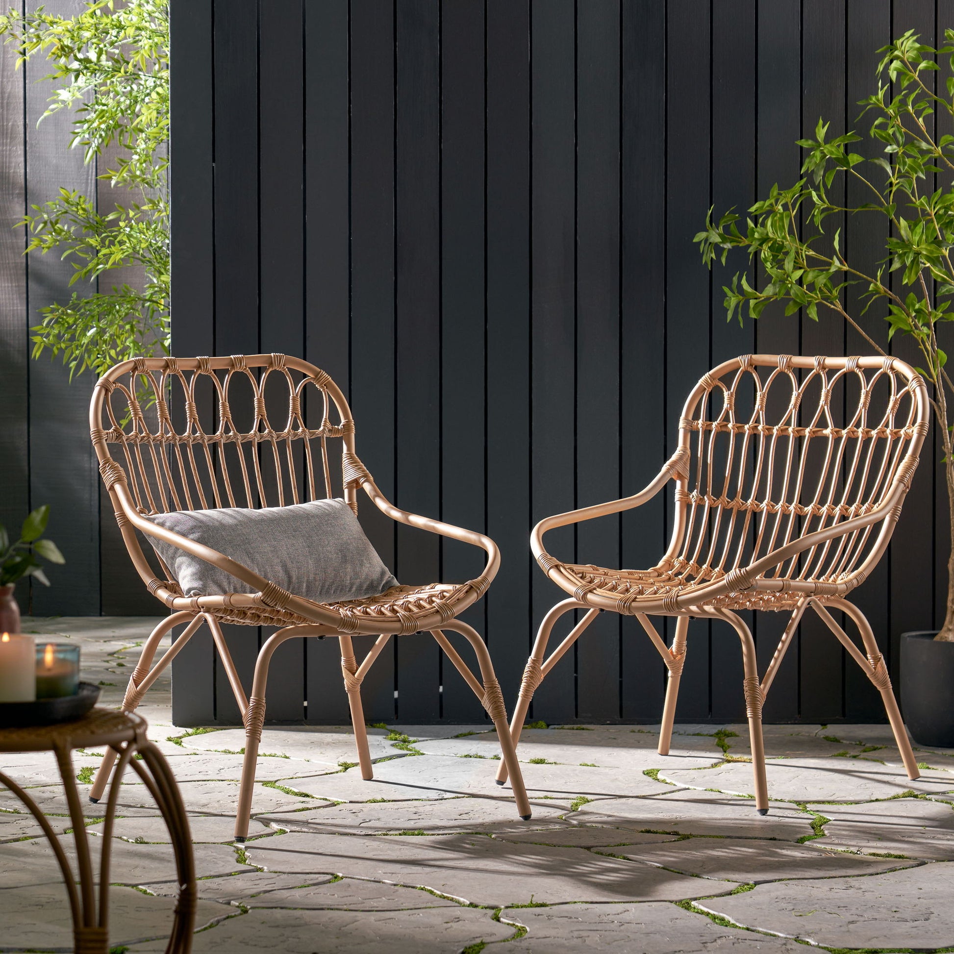 Two Monsoon Outdoor Accent Chairs with open weave design are arranged on a stone patio with a small table. One chair features a gray cushion. Greenery and a tall dark wooden fence complete the calm, inviting setting.