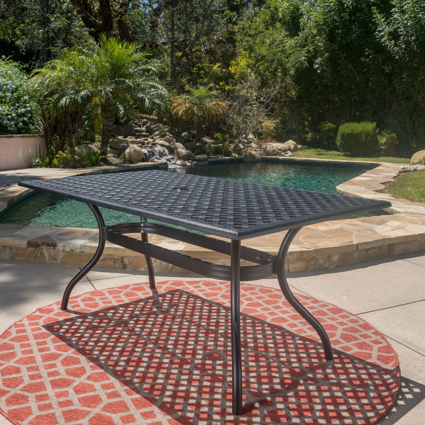 The Outdoor Aluminum Dining Table With Lattice Design And Center Umbrella Hole - Black sits on a red patterned rug by a swimming pool, surrounded by lush greenery in a sunny backyard.