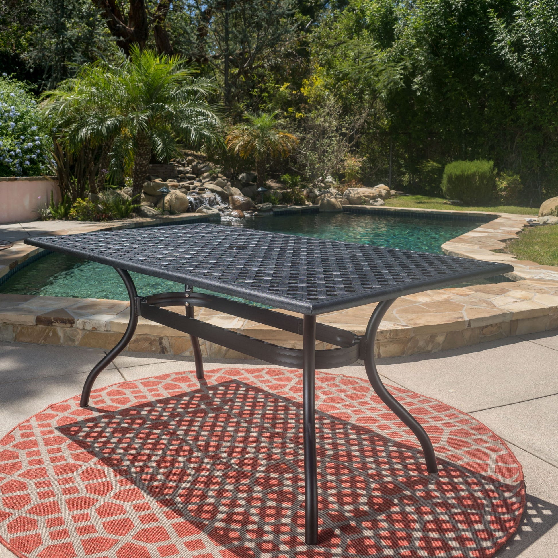The Outdoor Aluminum Dining Table With Lattice Design And Center Umbrella Hole - Black sits on a red patterned rug by a swimming pool, surrounded by lush greenery in a sunny backyard.