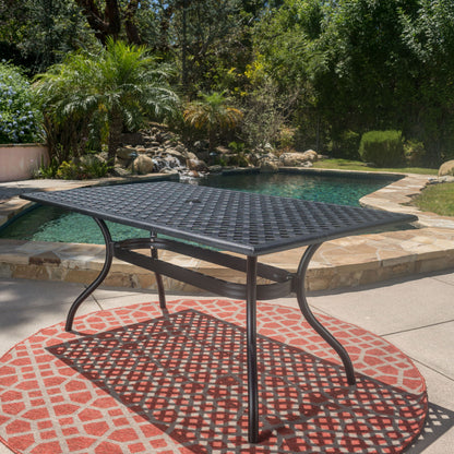 The Outdoor Aluminum Dining Table With Lattice Design And Center Umbrella Hole - Black sits on a red patterned rug by a swimming pool, surrounded by lush greenery in a sunny backyard.