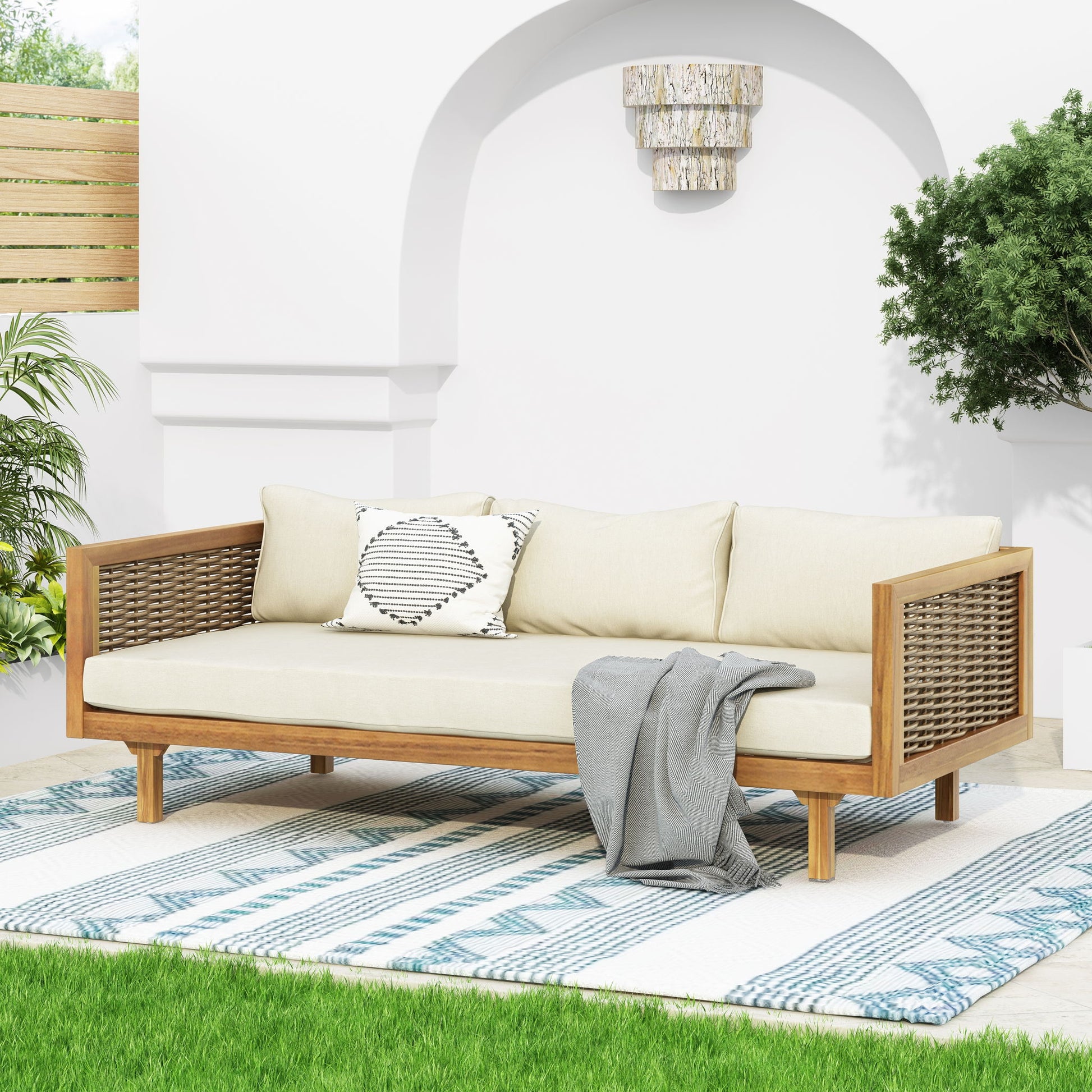 The Claremont 3 Seater Daybed, crafted from teak and acacia wood with water-resistant cream cushions, sits atop a blue and white patterned rug. A gray blanket and decorative pillow accent the seat, with lush green grass in the foreground.