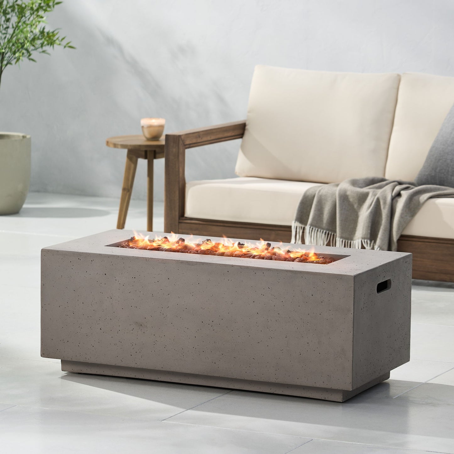 A Contemporary Outdoor 40,000 Btu Propane Fire Pit with flames is placed before a light outdoor sofa with wooden armrests and a gray throw, set on a patio featuring a potted plant in the background.