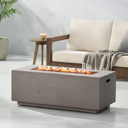 A Contemporary Outdoor 40,000 Btu Propane Fire Pit with flames is placed before a light outdoor sofa with wooden armrests and a gray throw, set on a patio featuring a potted plant in the background.