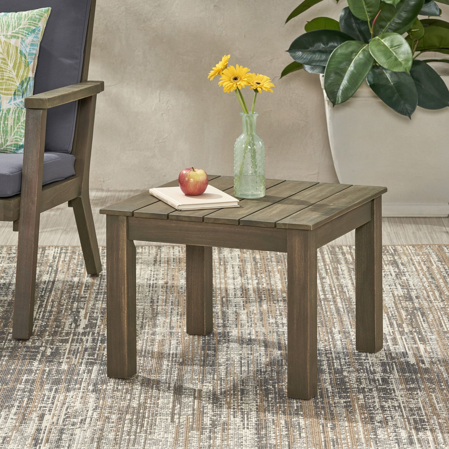 The Halcyon Outdoor Side Table with Slat Design holds a glass vase with yellow flowers, an apple, and a closed book. Positioned on a patterned rug, it sits beside an armchair and a large potted plant.