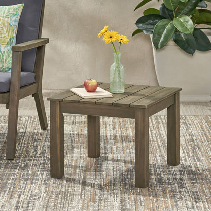 The Halcyon Outdoor Side Table with Slat Design holds a glass vase with yellow flowers, an apple, and a closed book. Positioned on a patterned rug, it sits beside an armchair and a large potted plant.