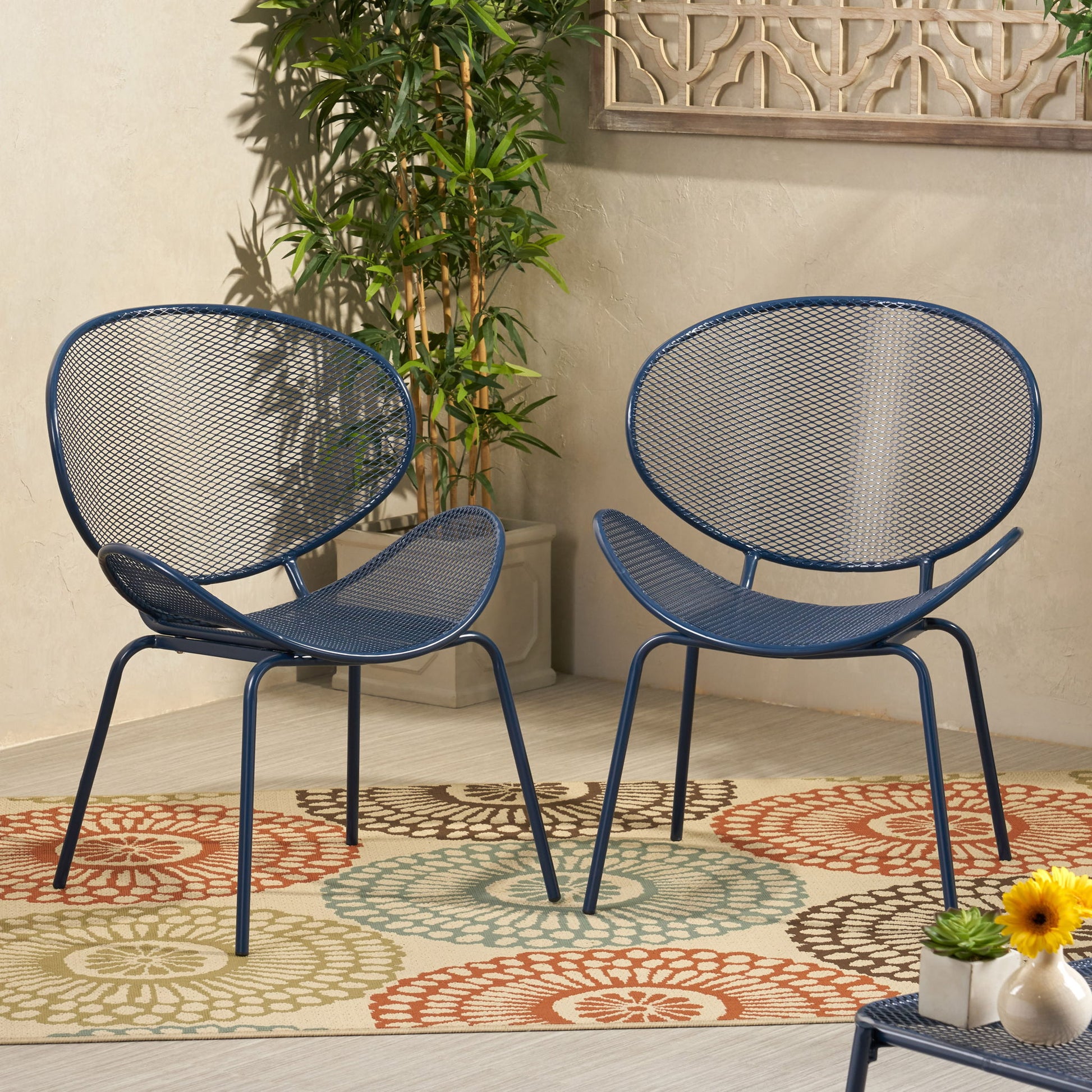 The Elba Contemporary Chair Set for Outdoor Relaxation features two modern blue metal chairs with mesh backs and seats, styled on a circular-patterned rug, with a small floral table, a green potted plant, and a decorative wall panel nearby.