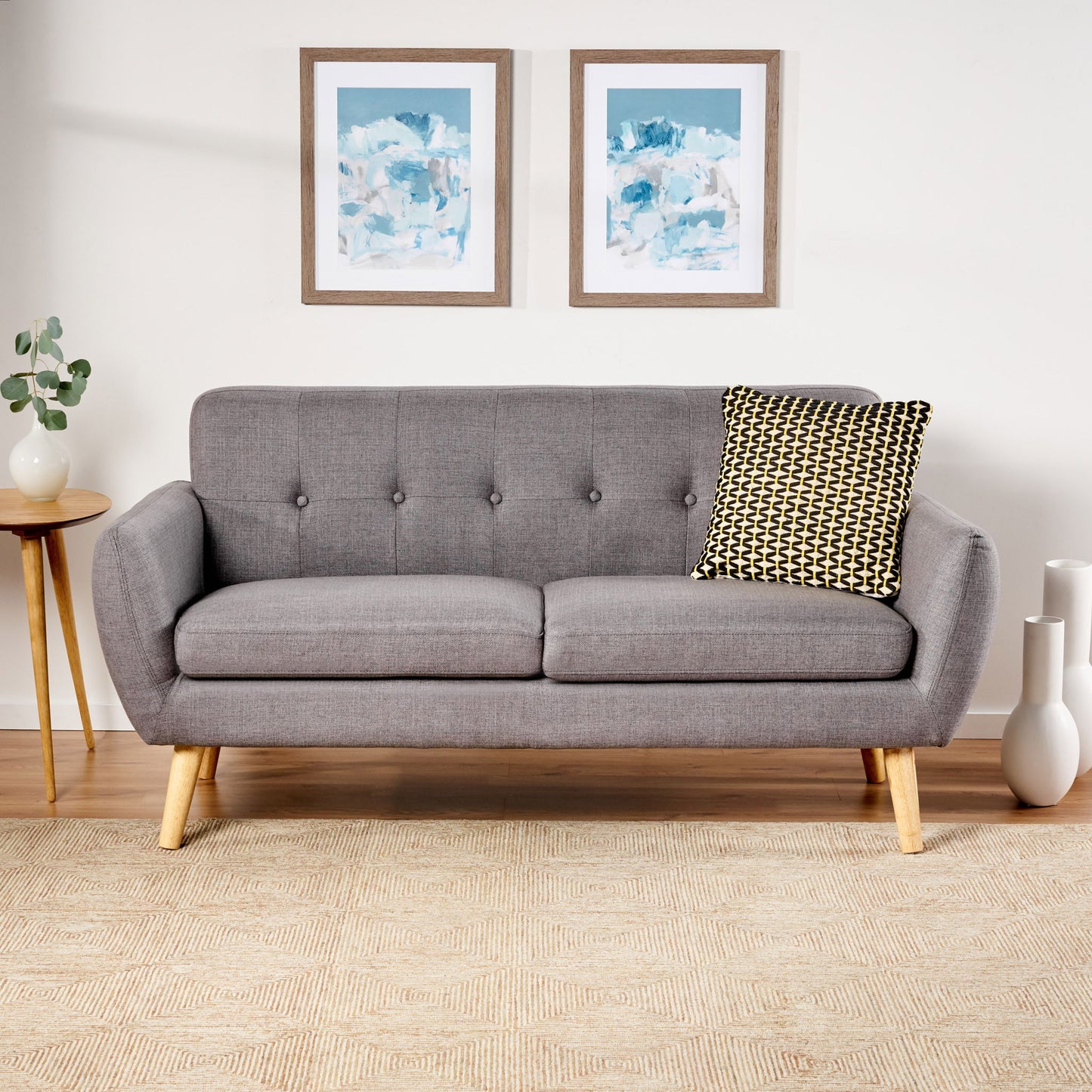 A Modern Loveseat Sofa With Button Tufted Backrest and wooden legs sits on a beige patterned rug, accented by a black and white pillow. Two abstract framed artworks hang above, with a small side table and plant to the left.