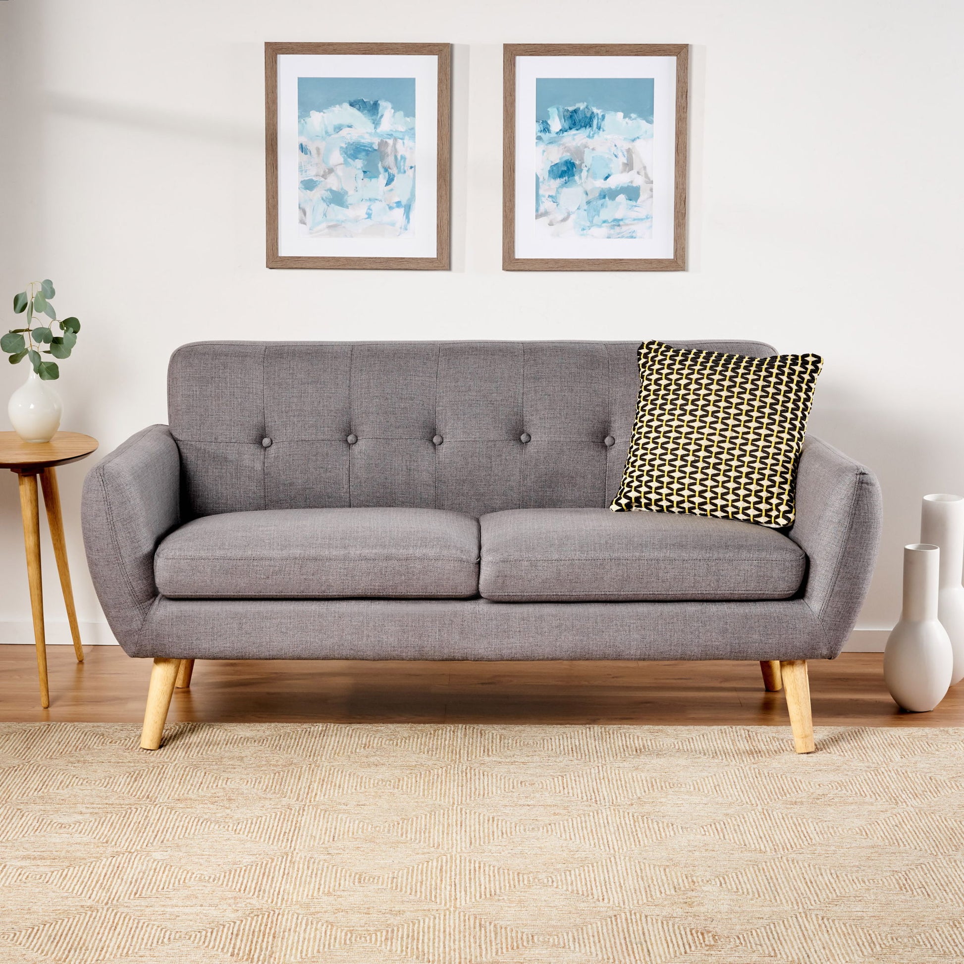 A Modern Loveseat Sofa With Button Tufted Backrest and wooden legs sits on a beige patterned rug, accented by a black and white pillow. Two abstract framed artworks hang above, with a small side table and plant to the left.
