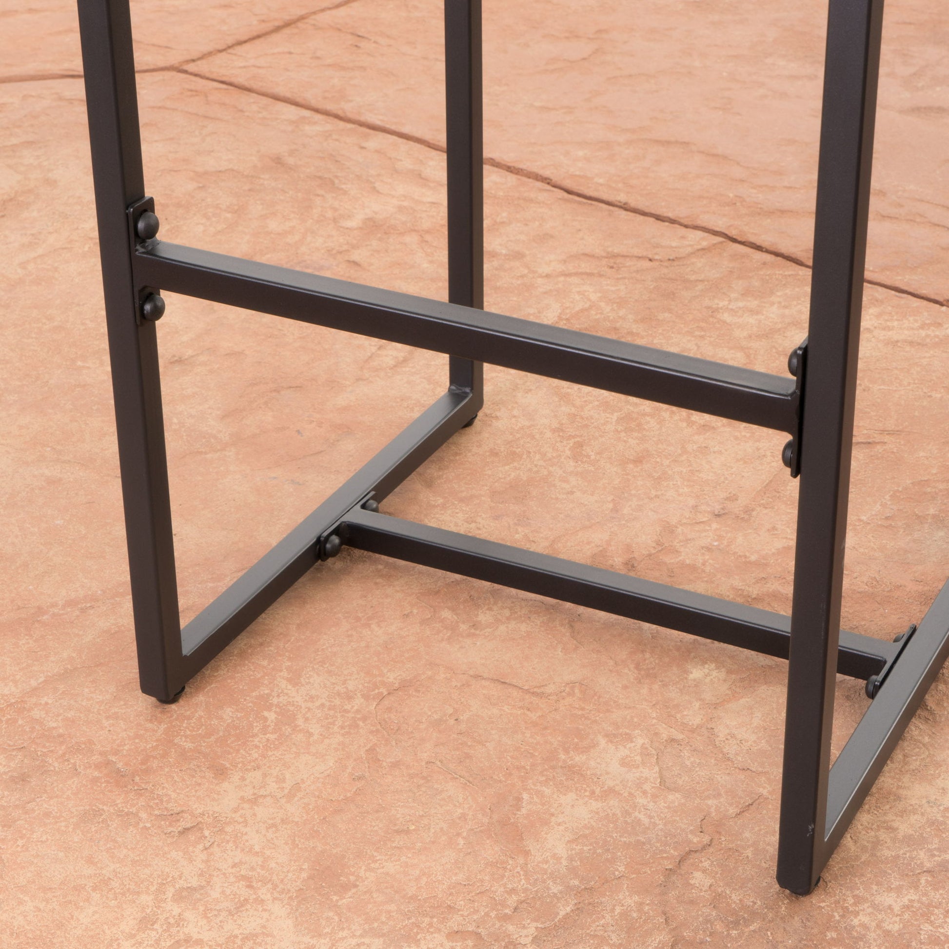 Close-up of the metal legs and lower frame of the Outdoor Barstool With Hand Crafted Details (Set of 2), shown on a textured reddish-brown stone patio.
