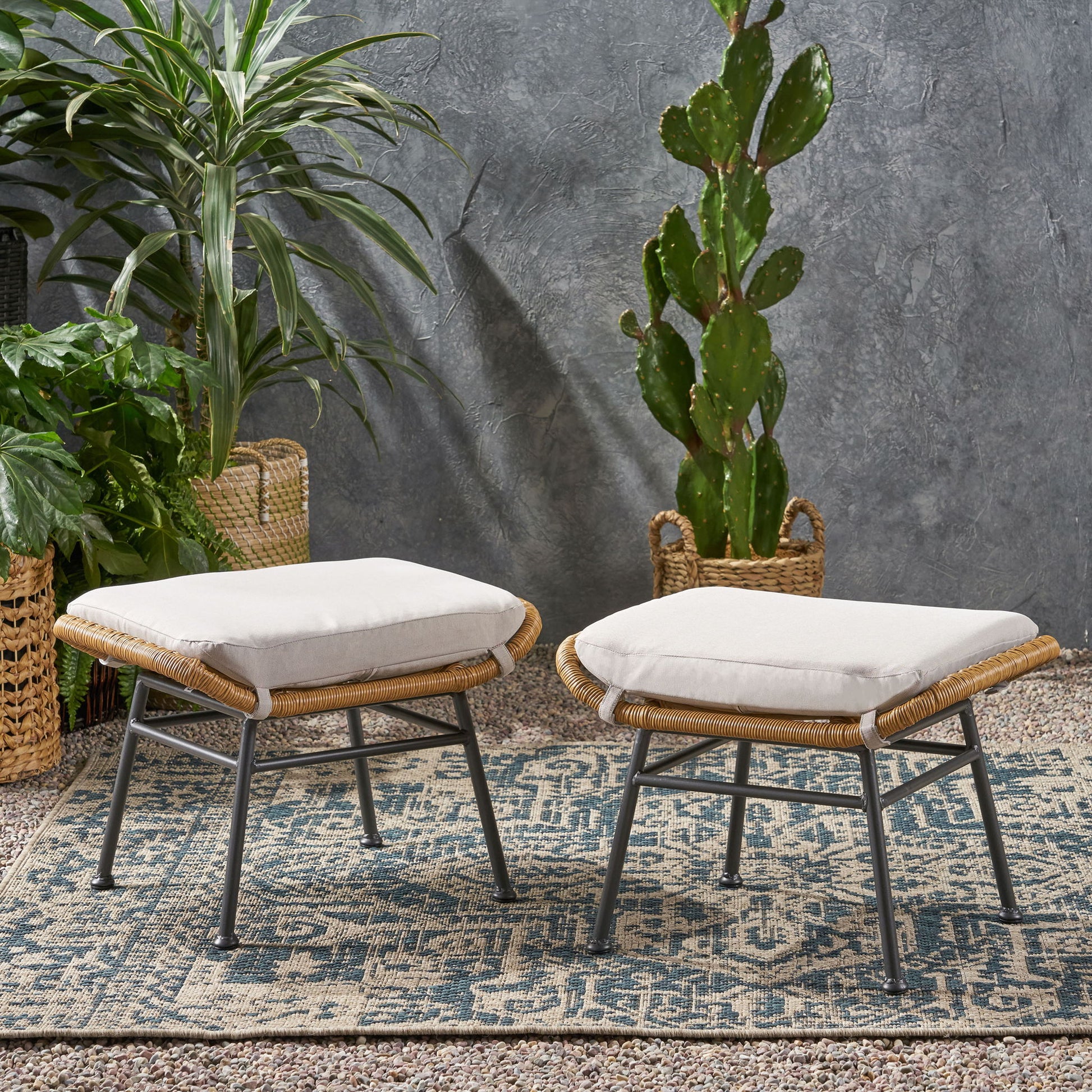 A pair of Outdoor Modern Boho Wicker Ottomans With Cushions are set on a patterned rug, accented by potted green plants and a textured gray wall.