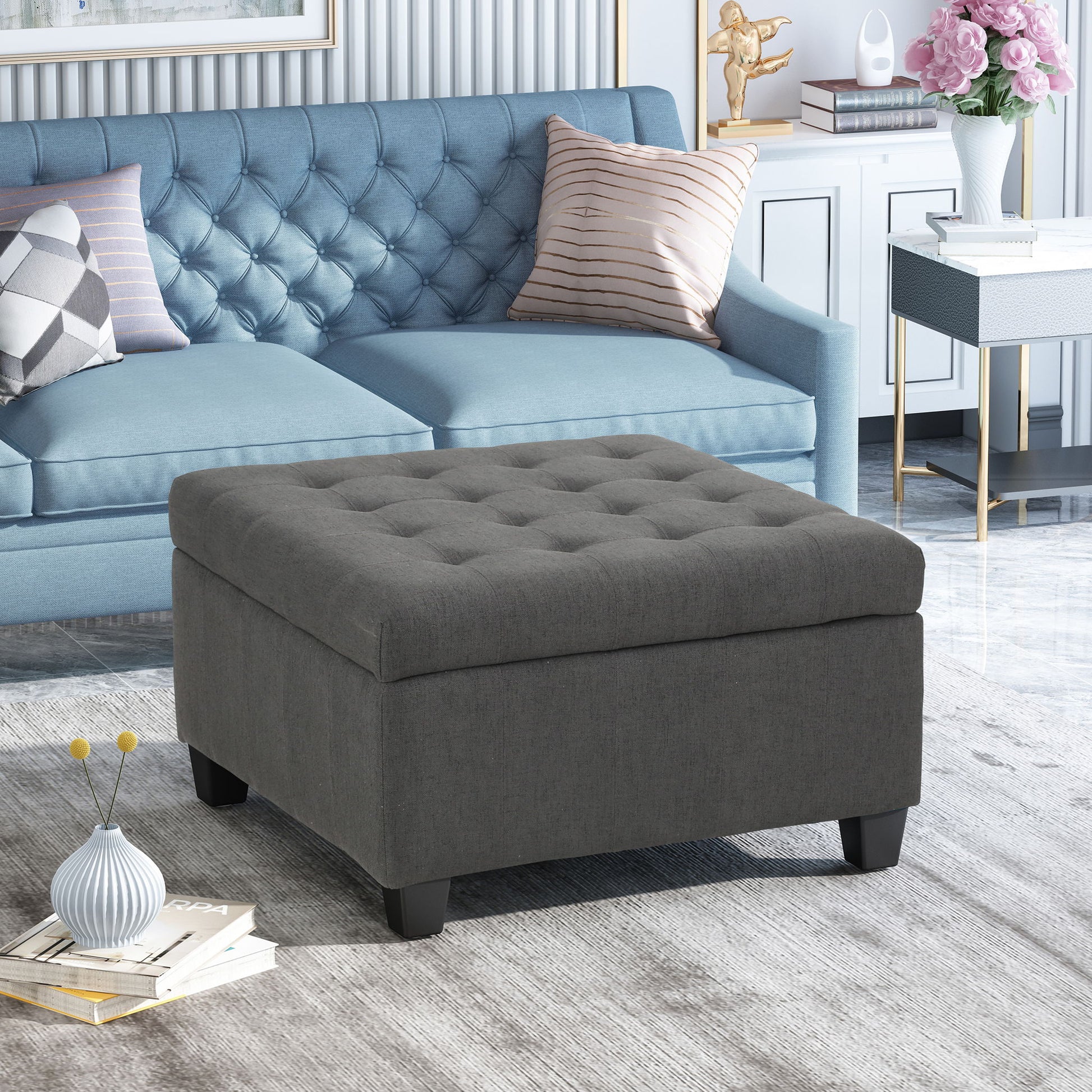 A modern living room with a blue tufted sofa, the Isabella - Contemporary Ottoman With Storage in gray, a white cabinet with pink flowers, decorative items, books on a light rug, and marble flooring.