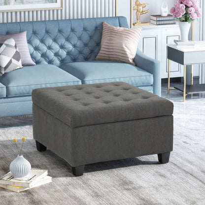 A modern living room with a blue tufted sofa, the Isabella - Contemporary Ottoman With Storage in gray, a white cabinet with pink flowers, decorative items, books on a light rug, and marble flooring.