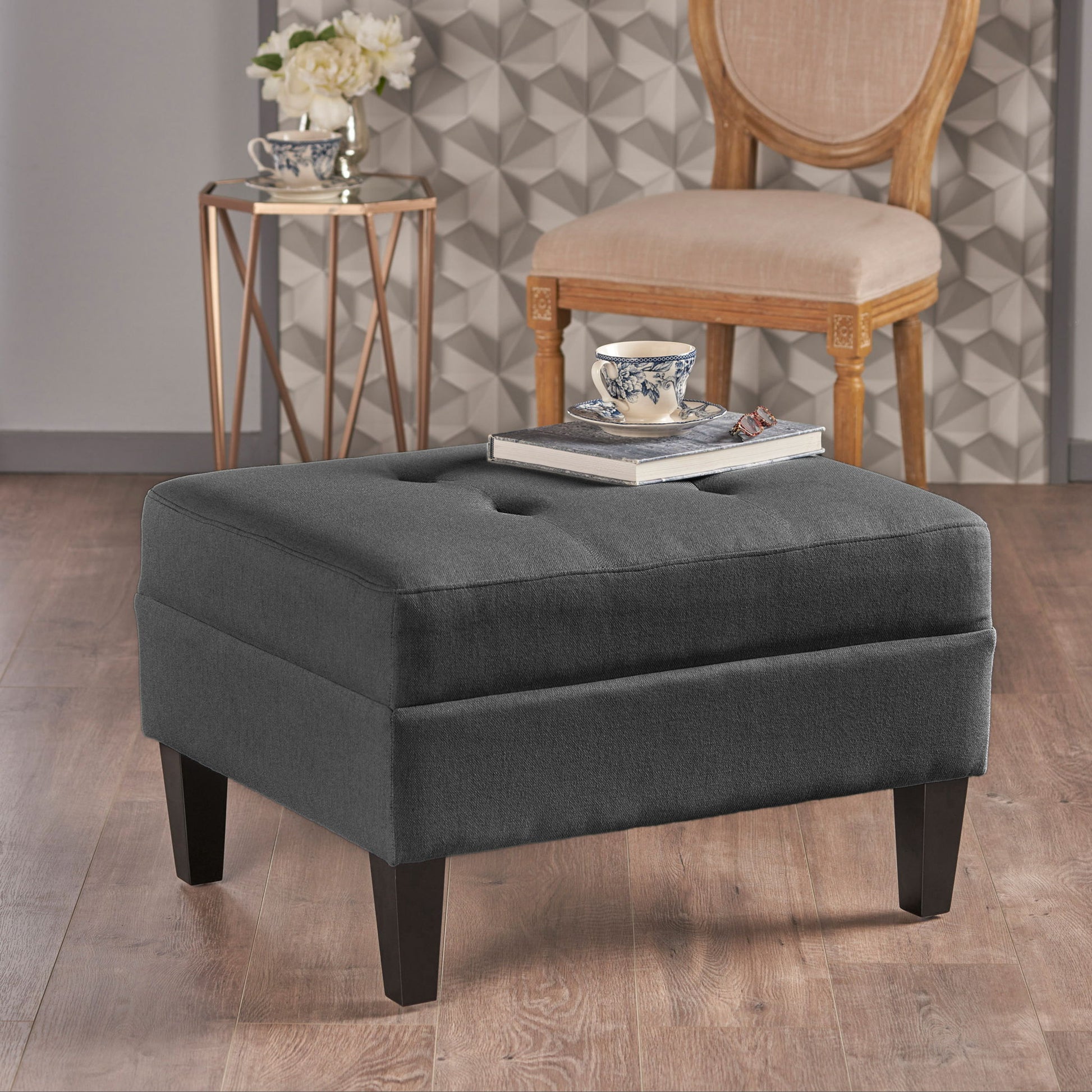 A Classic Ottoman With Birch Wood Legs And Fabric Upholstery sits on a wooden floor with books and a teacup on top. Behind it are a beige chair, side table with flowers, and a wall featuring a geometric pattern.