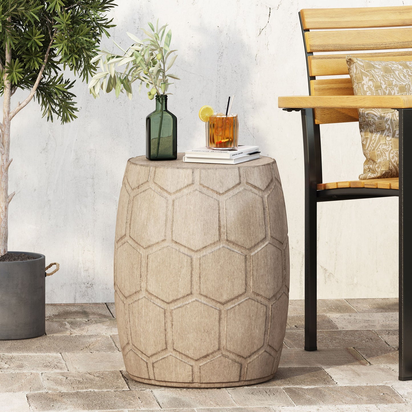 An Outdoor Lightweight Concrete Side Table in Natural holds a green glass vase with olive branches and a cocktail by a wooden bench on a patio with a potted tree.