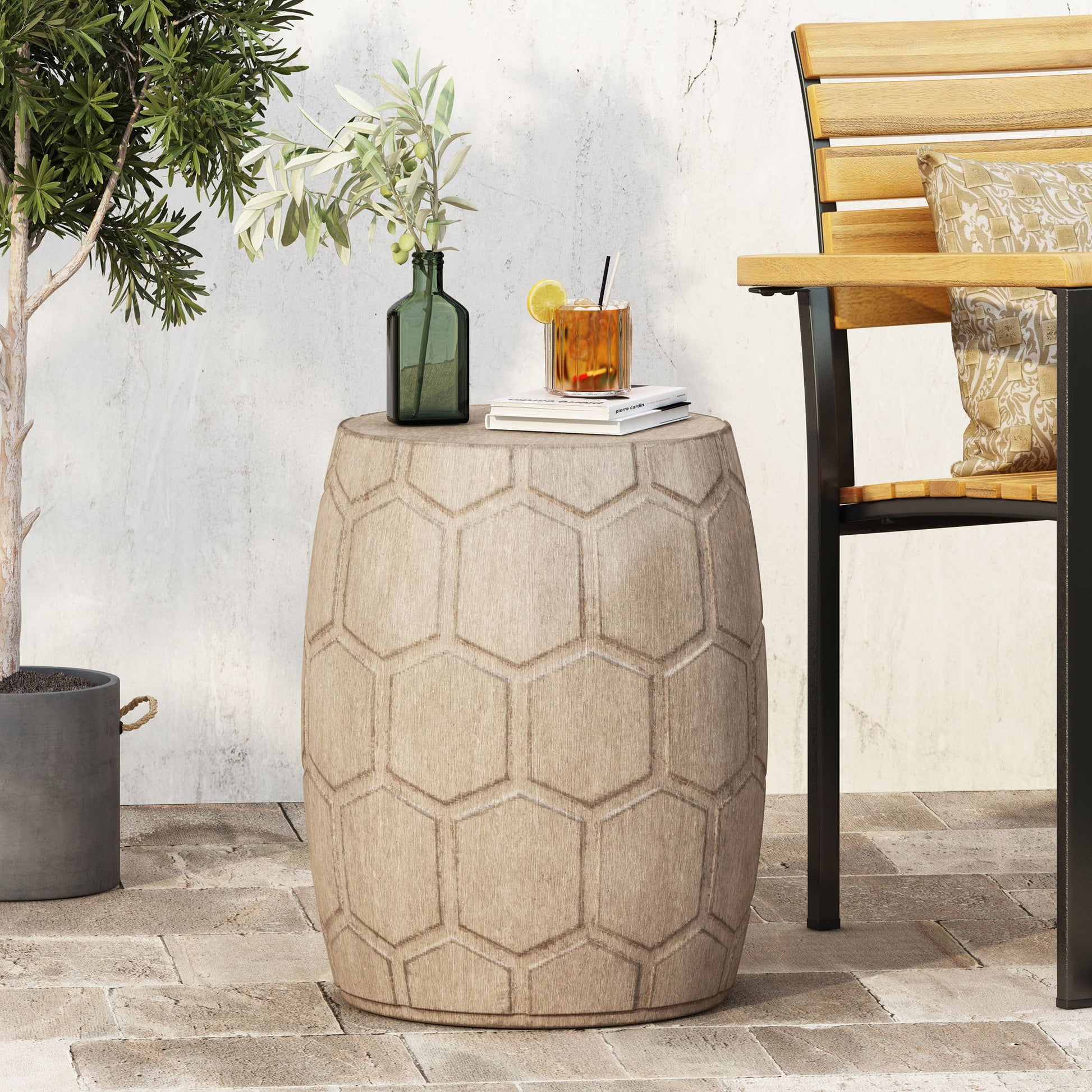 An Outdoor Lightweight Concrete Side Table in Natural holds a green glass vase with olive branches and a cocktail by a wooden bench on a patio with a potted tree.