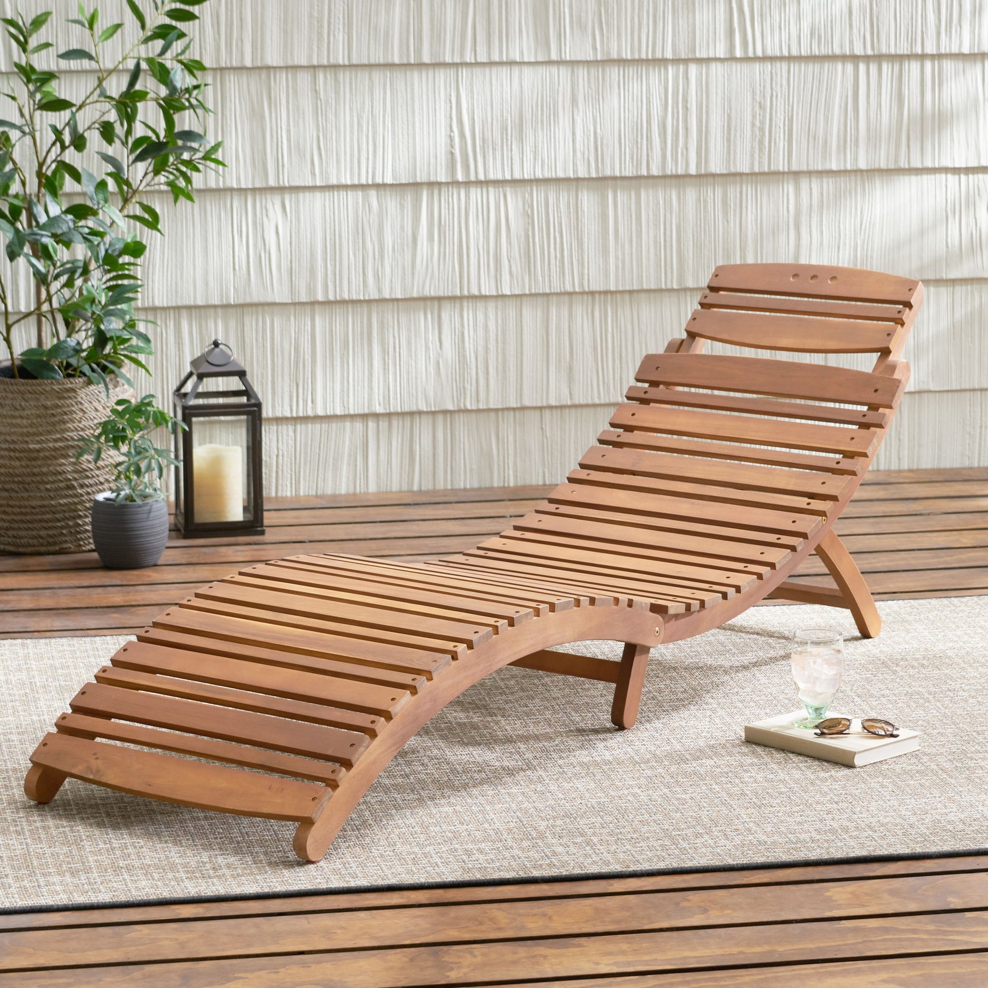 The Lahaina - Wood Foldable Chaise Lounge rests on an outdoor rug atop a wooden deck, accompanied by a potted plant, candle lantern, drink, and open book beside a shiplap wall.