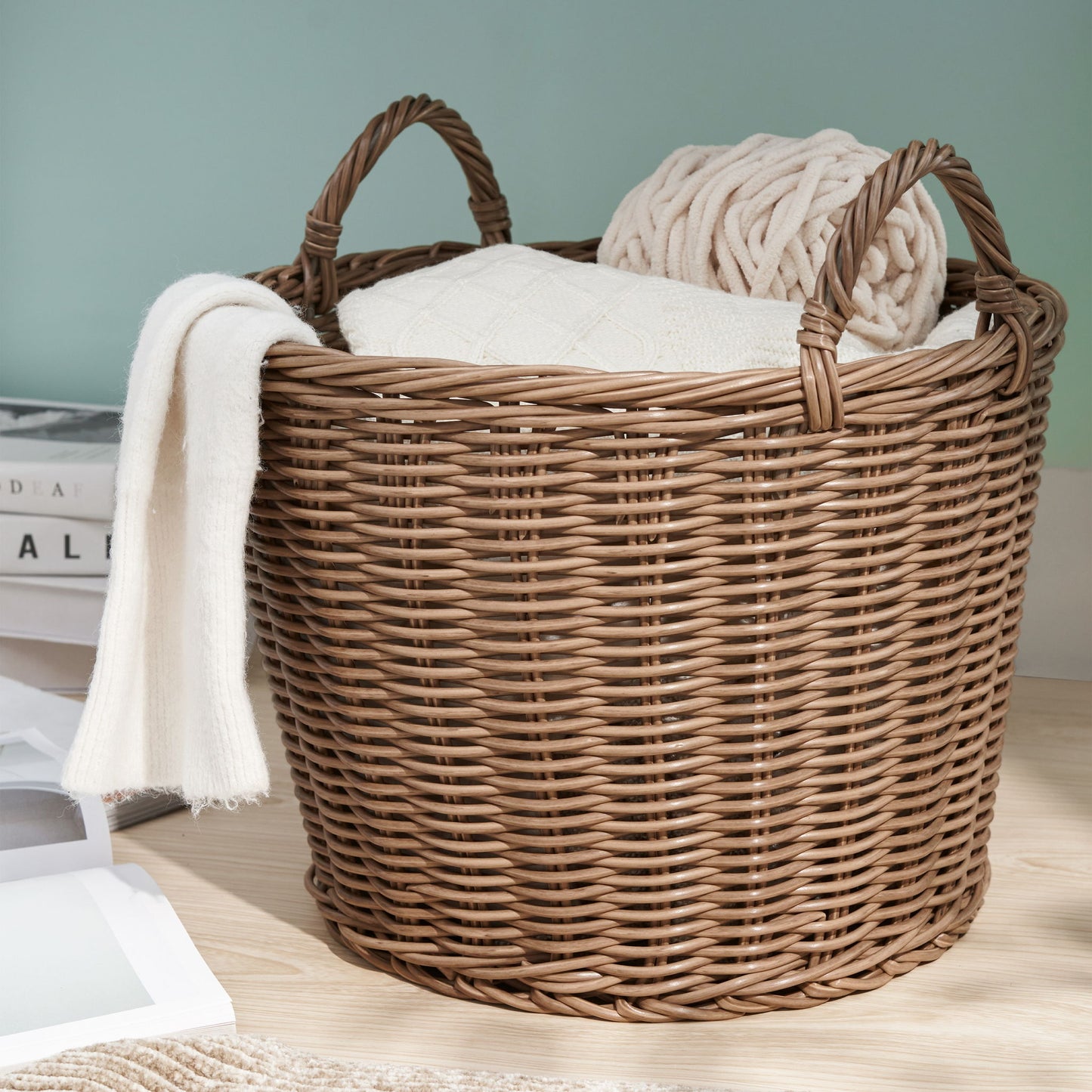 A Round Cone Woven Resin Wicker Basket With Handles in walnut brown sits on a wooden floor, holding a folded white blanket and beige knitted throw. Nearby are a stack of books and a white picture frame.