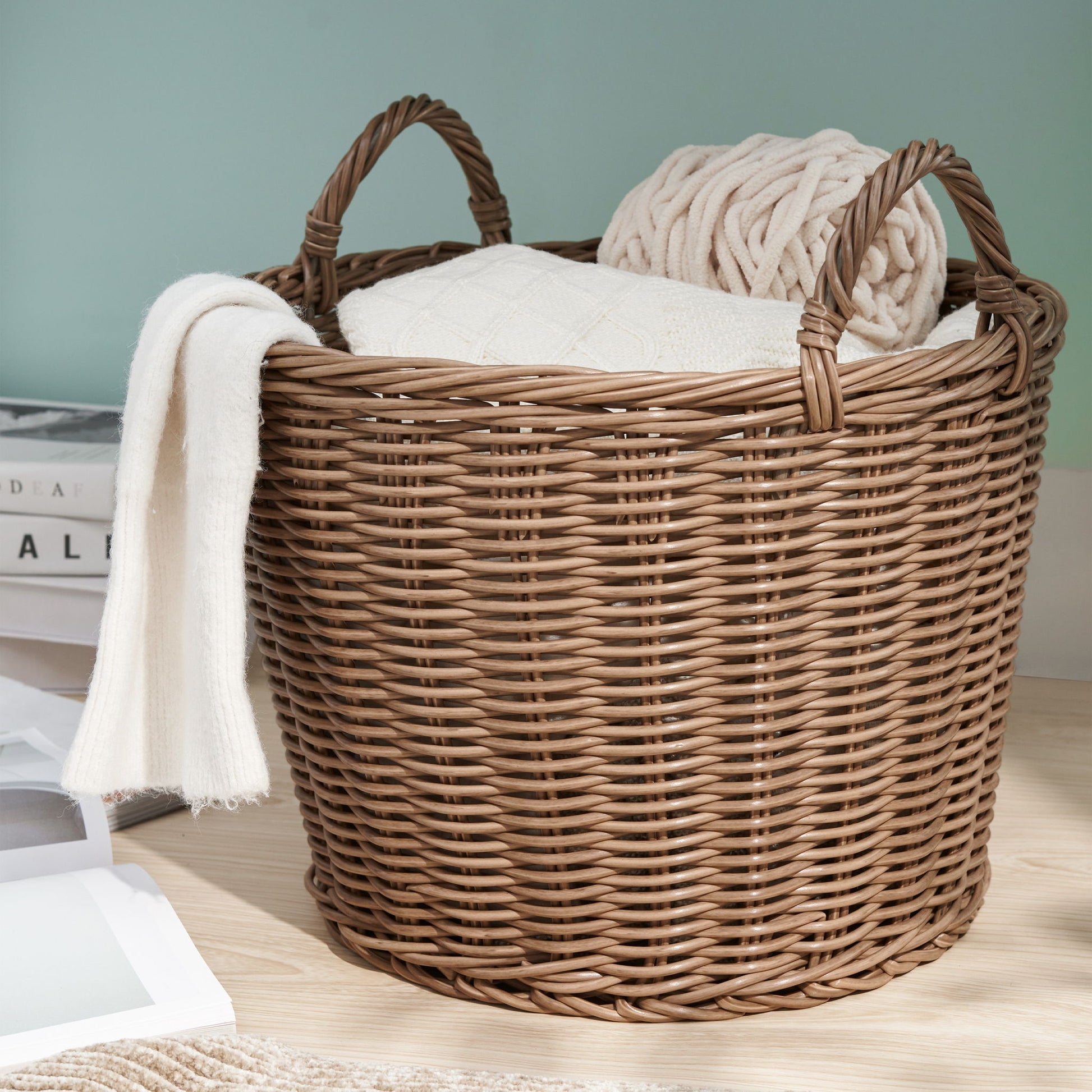 A Round Cone Woven Resin Wicker Basket With Handles in walnut brown sits on a wooden floor, holding a folded white blanket and beige knitted throw. Nearby are a stack of books and a white picture frame.